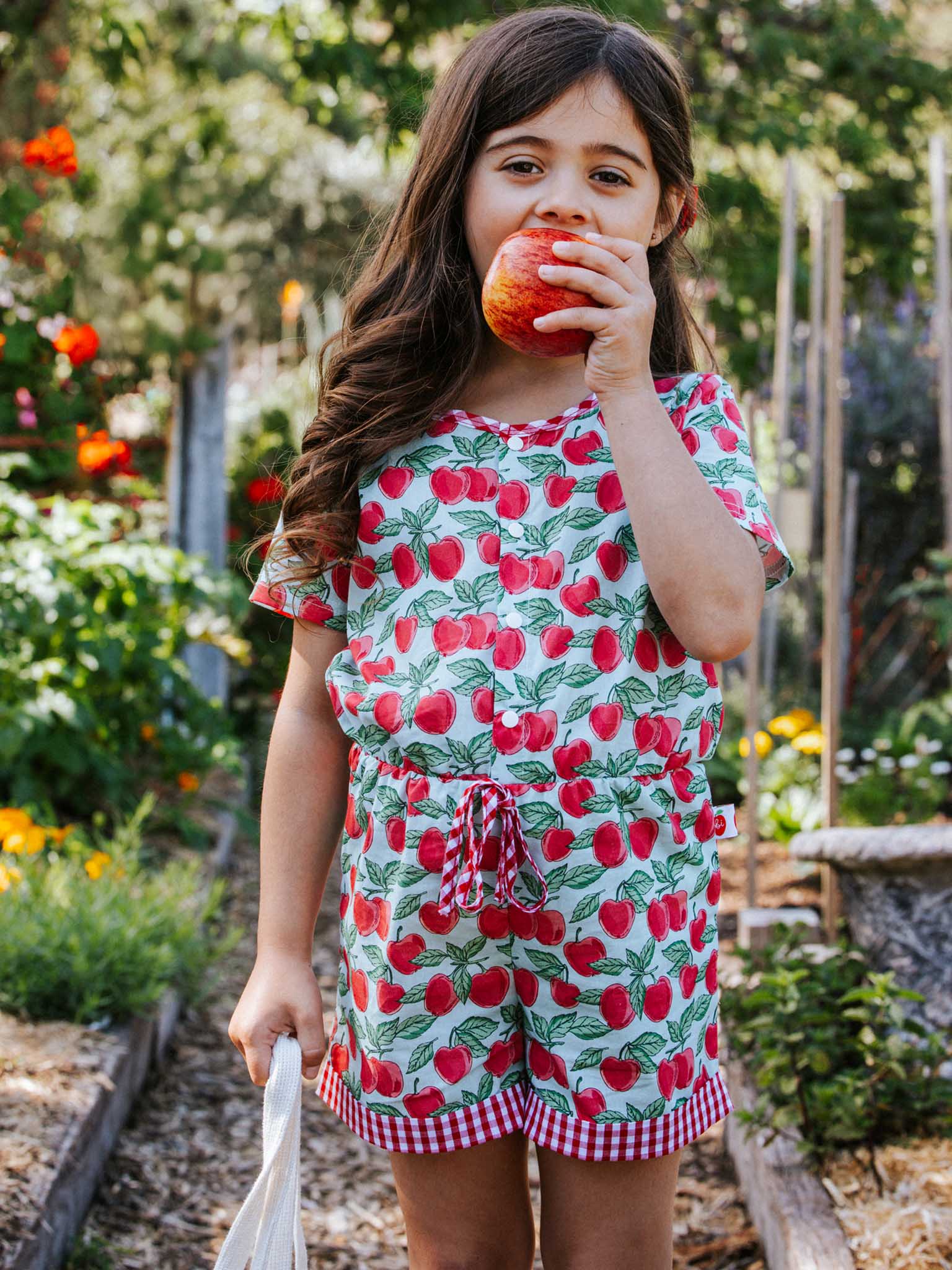 A young girl in a Blue Cherry Maisy Playsuit stands outdoors, holding an apple to her mouth, surrounded by greenery and flowers.