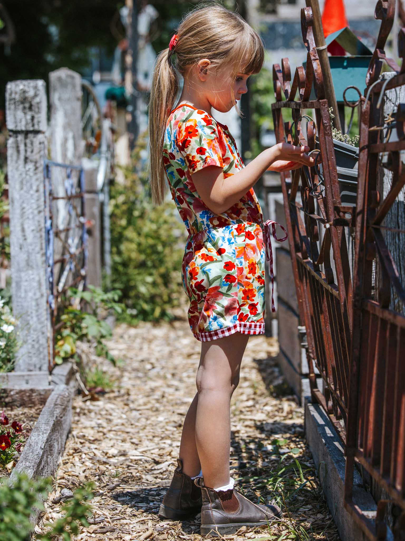 A young girl in the vibrant Wild Roses Maisy Jersey Playsuit stands by a metal fence in a garden, observing something closely.