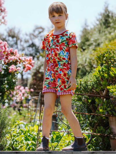 A young child stands in a garden wearing a Wild Roses Maisy Jersey Playsuit paired with boots.