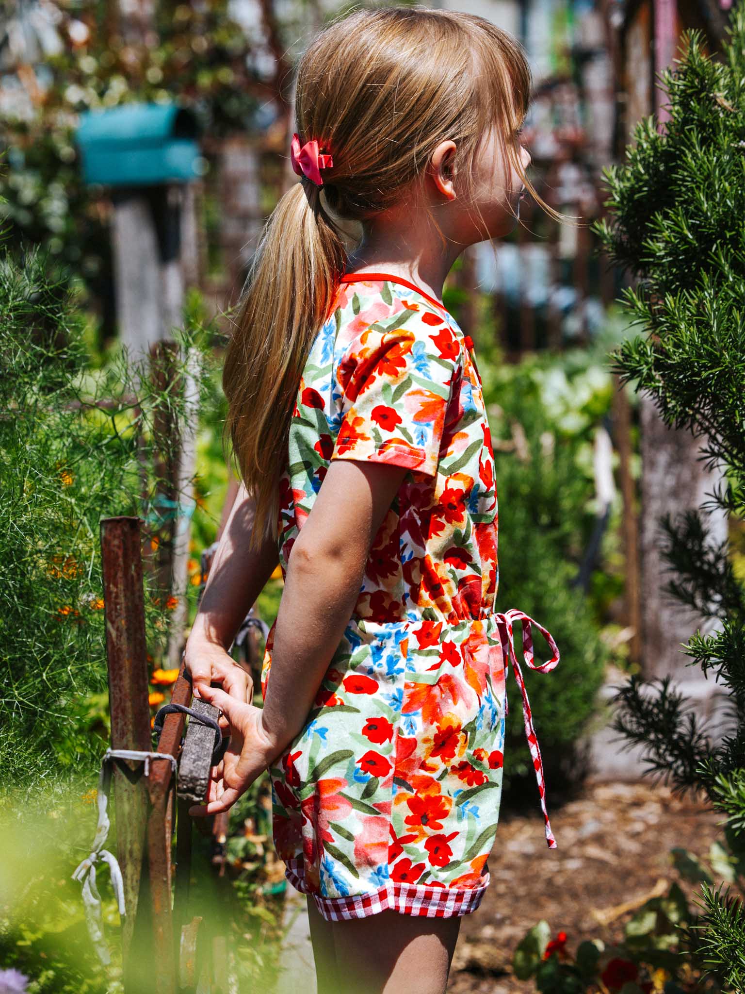 In a vibrant garden, a young girl wearing the delightful Wild Roses Maisy Jersey Playsuit gazes to the side, her ponytail swaying gently. Holding onto a small gate enveloped in lush greenery, she perfectly captures the carefree essence of summer highlighted in Wild Roses' playful girls' clothing collections.