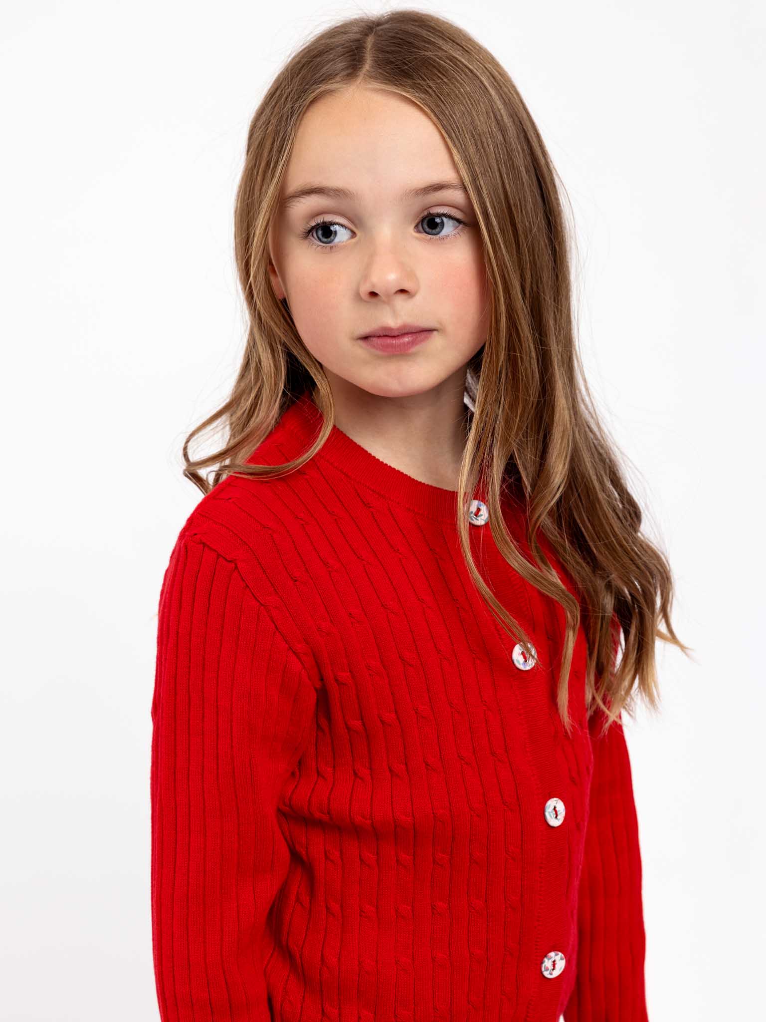 A young girl with long hair wears a Knitwear Maxine Red Cardigan, featuring fabric-covered buttons, and looks to the side against a plain white background.