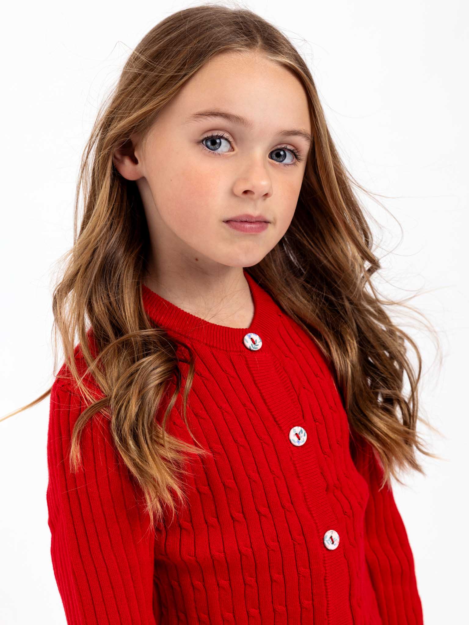 A young girl with long brown hair, wearing a Maxine Red Cardigan by Knitwear, which features a cotton cable knit and charming fabric-covered buttons, gazes at the camera against a plain white background.