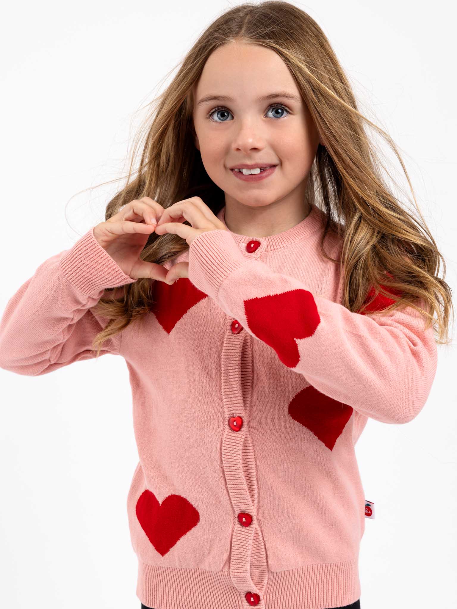 A girl with long hair wears a Knitwear Maxine Pink with Red Hearts Cardigan, showcasing its heart-shaped buttons, and forms a heart shape with her hands against a plain background.