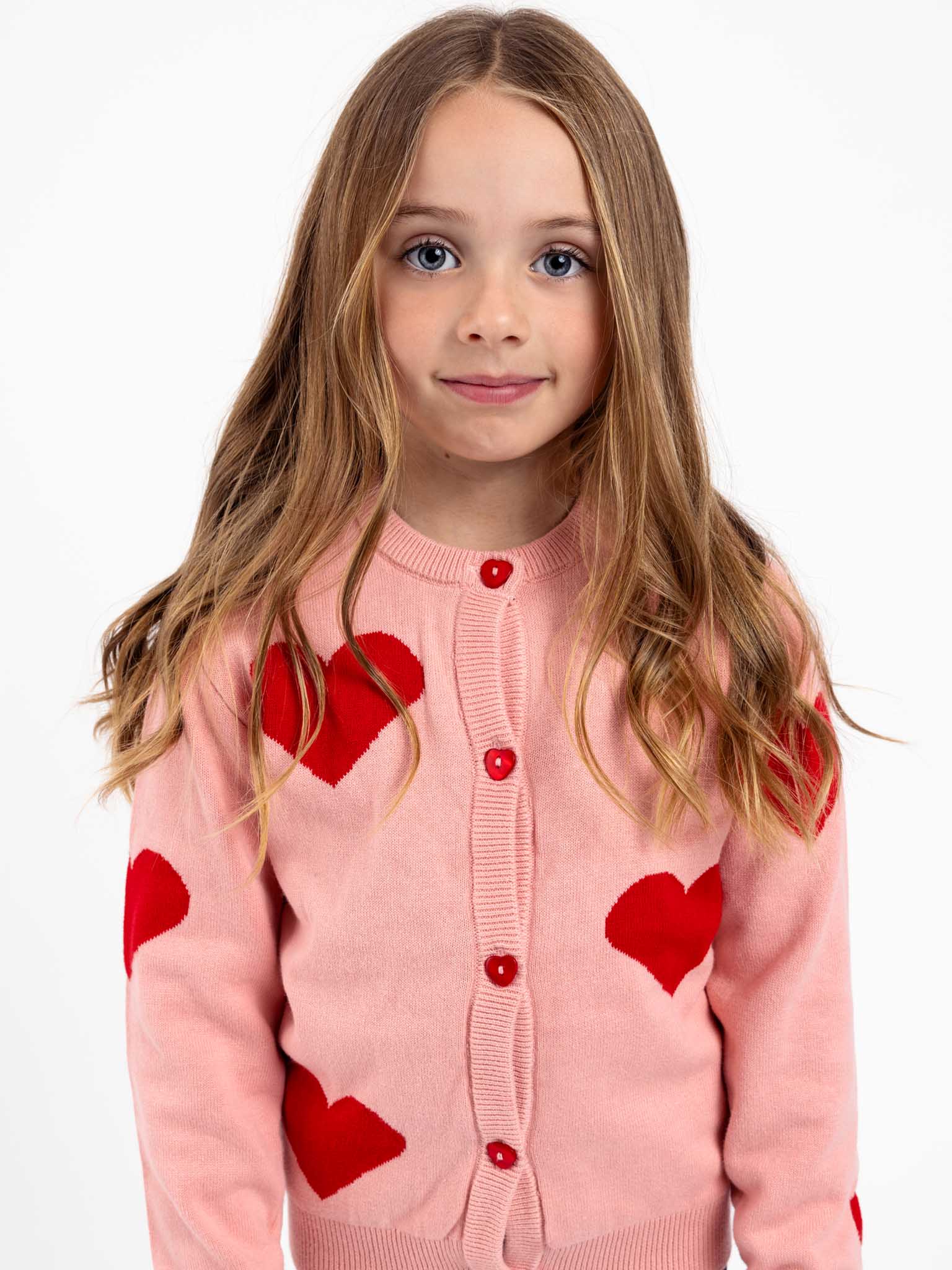 A young girl with long hair is wearing a Knitwear Maxine Pink with Red Hearts Cardigan, standing against a white background.