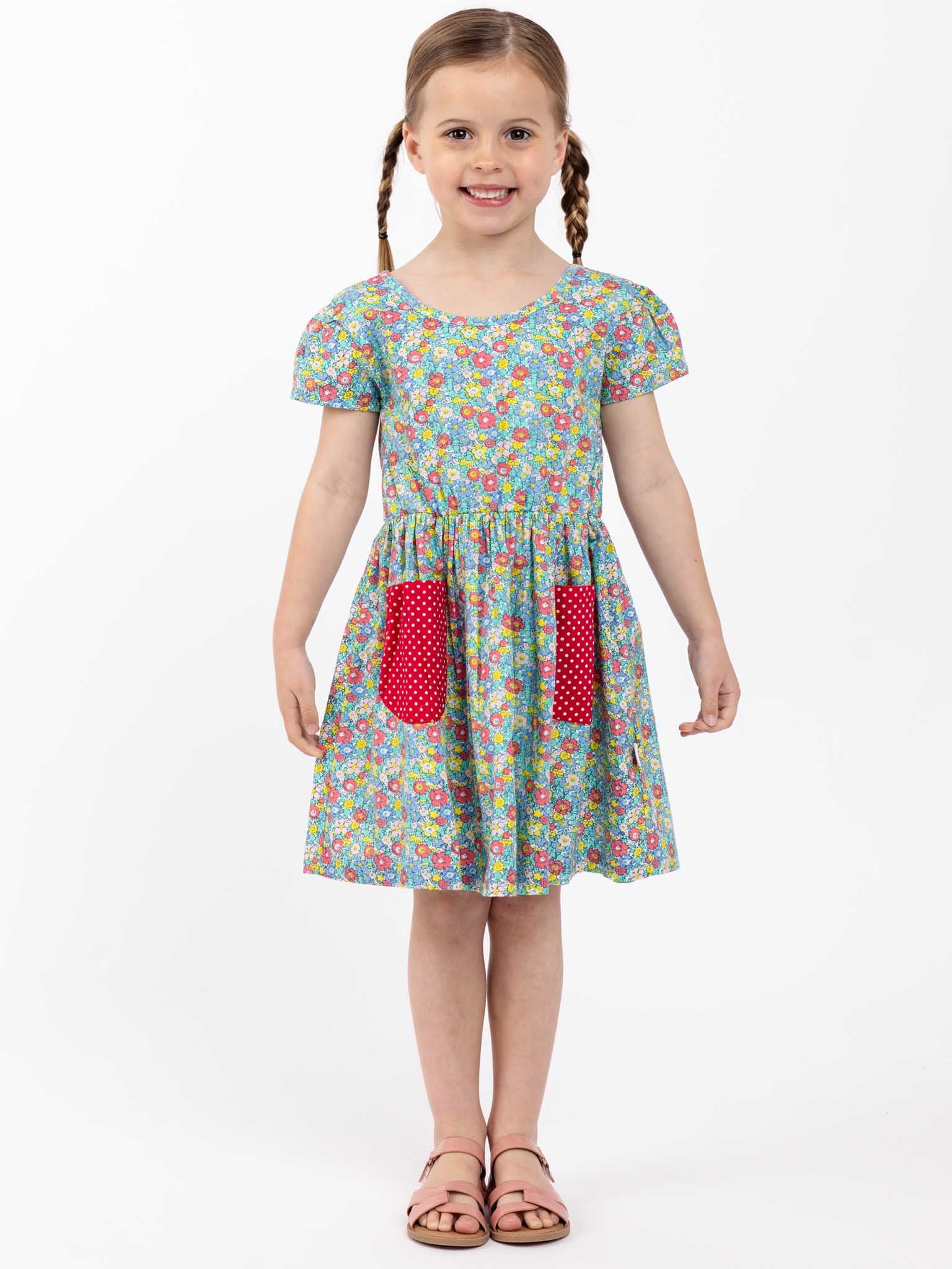 A little girl in a Sweetie Bows Minifloral Dress by New Minifloral, standing on a white background, exuding cuteness.