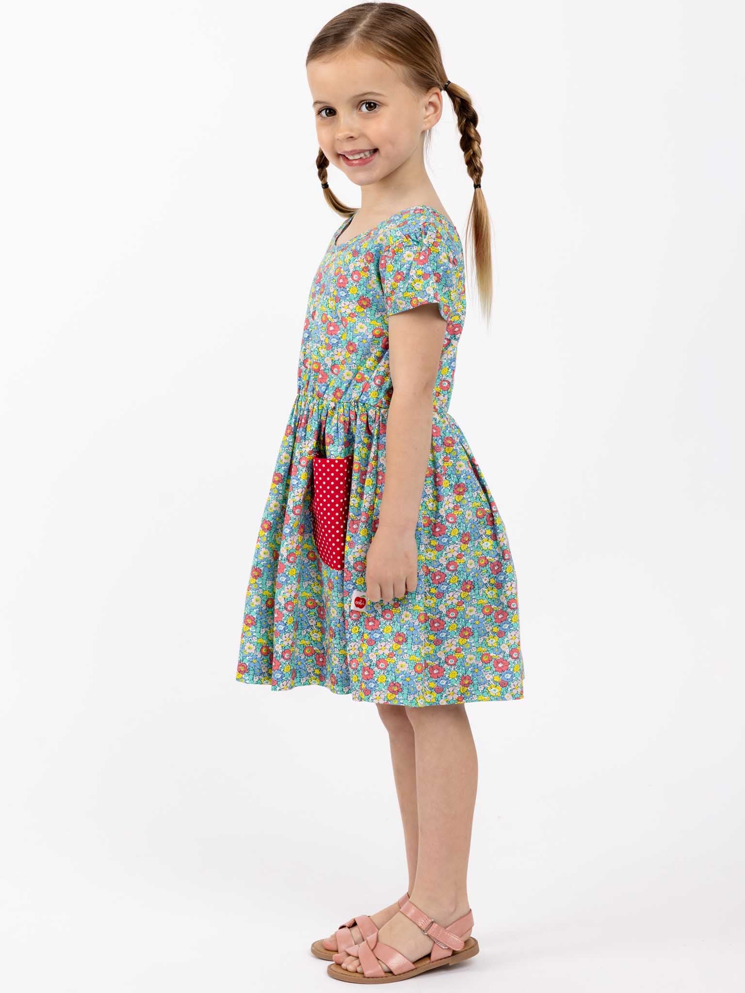 A little girl in a Sweetie Bows Minifloral Dress from New Minifloral, standing on a white background, showcasing cuteness overload.