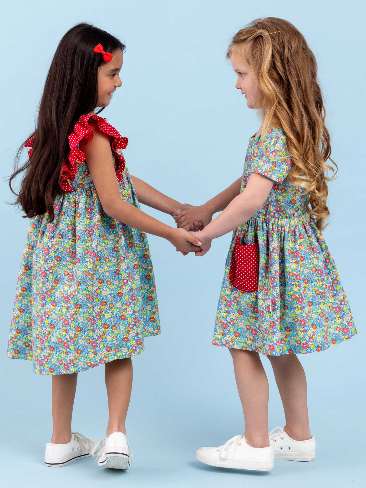 Two little girls in ruffle sleeves are holding hands in front of a blue background wearing the Molly Minifloral Dress by New Minifloral.