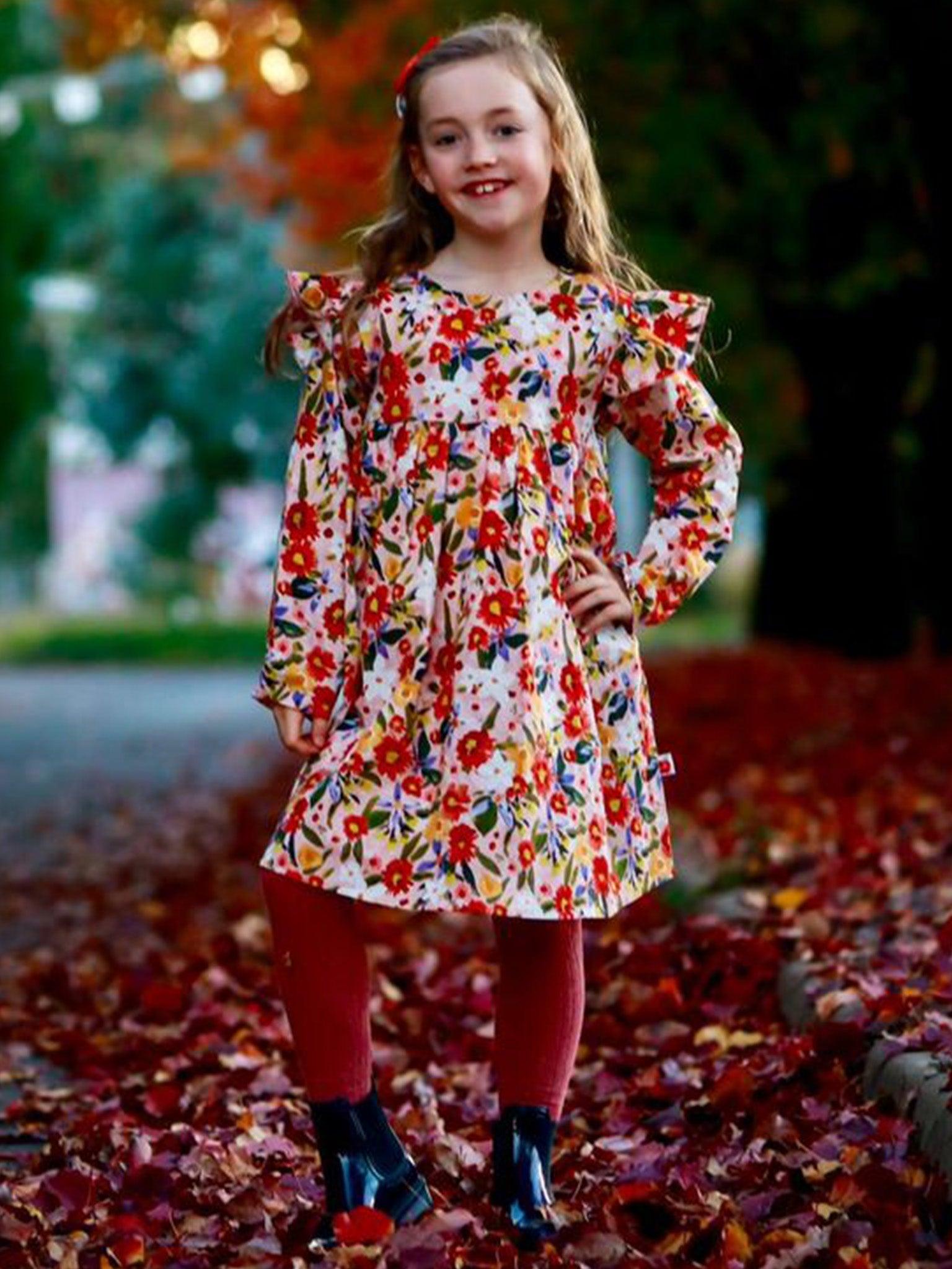 A young girl stands on a leaf-strewn path, wearing a Native Bloom dress in earthy colours, red tights, black shoes, and smiling at the camera. The background features autumn foliage and blurred trees, enhancing her natural beauty.