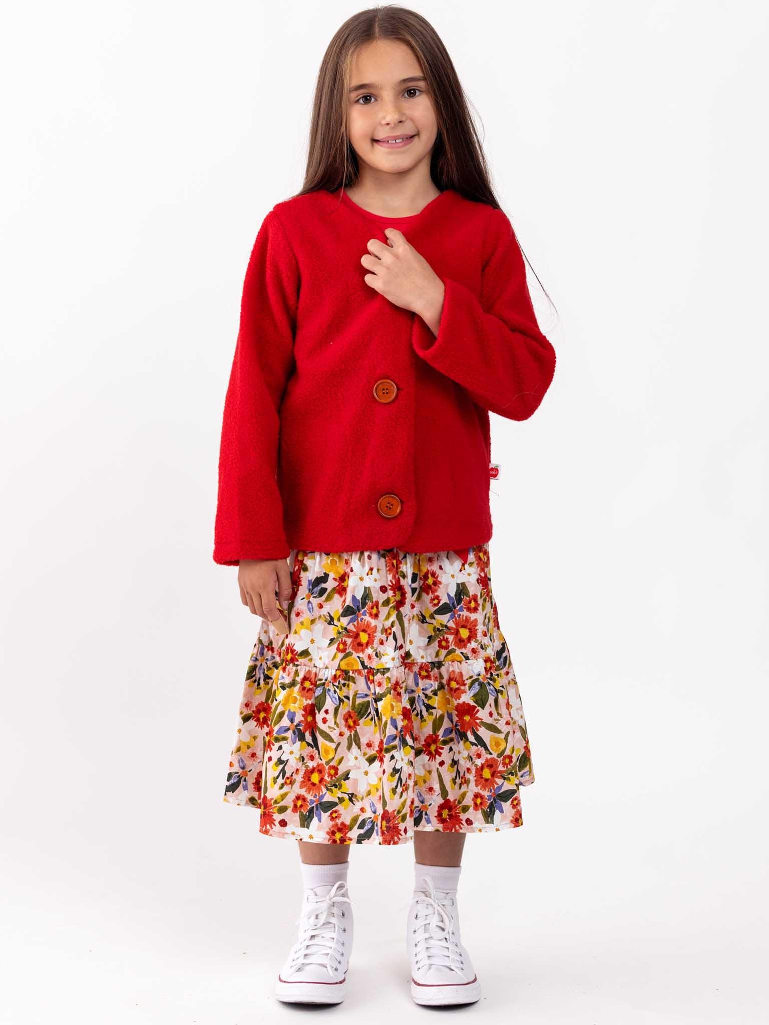 A young girl in a red cardigan and a Native Bloom maxi skirt standing against a white background, smiling gently.