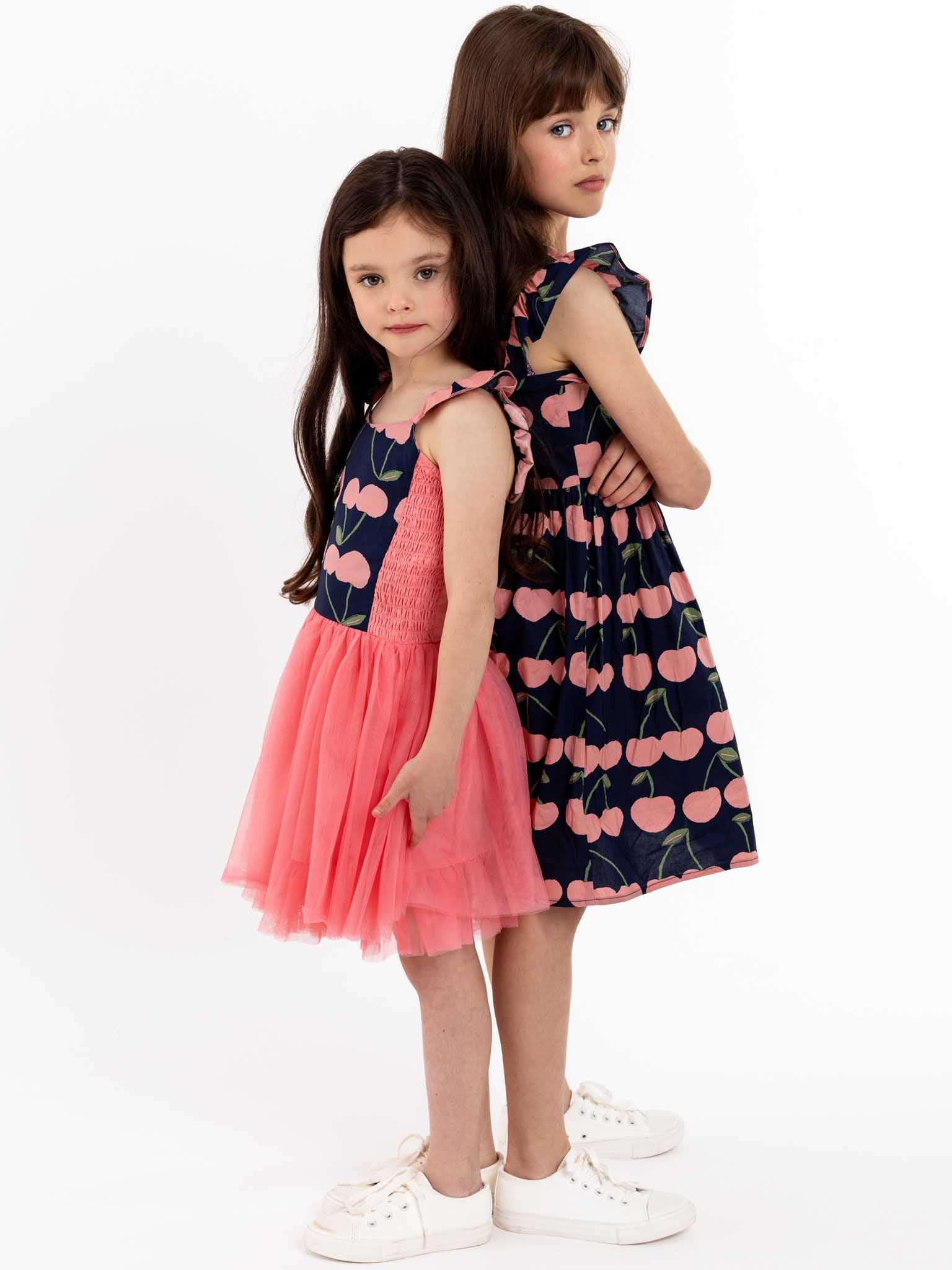 Two little girls in pink and blue dresses standing next to each other, showcasing their summer outfits with adorable Navy Cherry Calista Dresses.