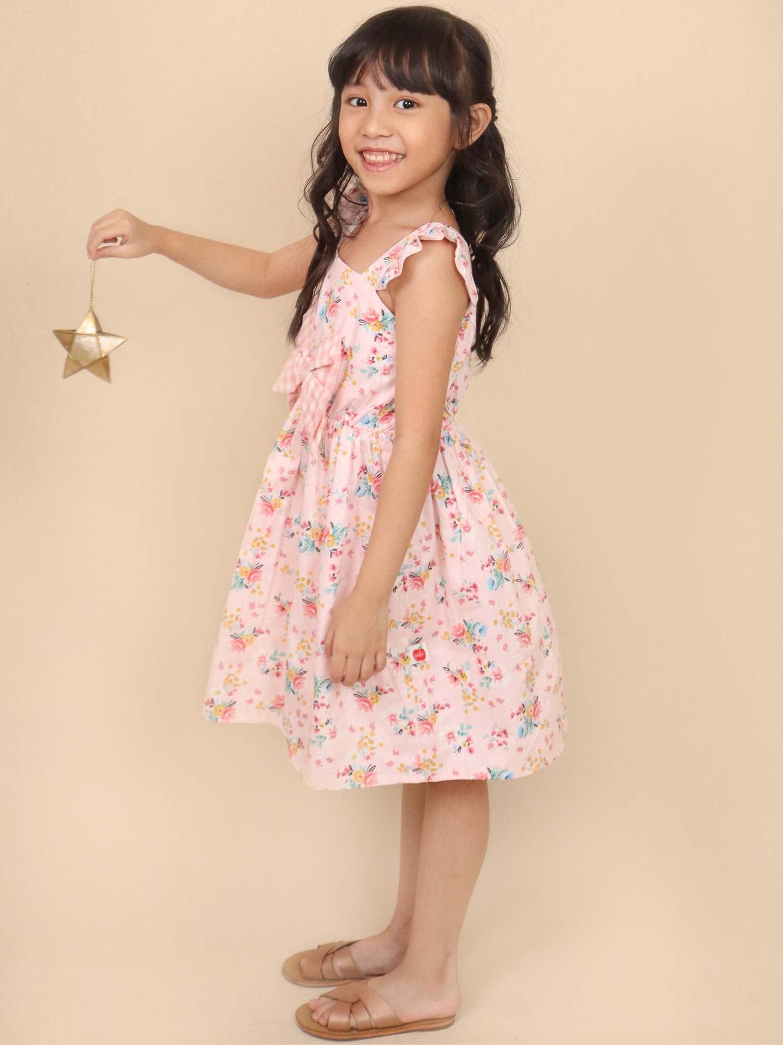 A young girl wearing the Vintage Pink Floral Mia Dress by Pink Flowers smiles sweetly as she holds a small gold star ornament against a beige background, exuding vintage charm.