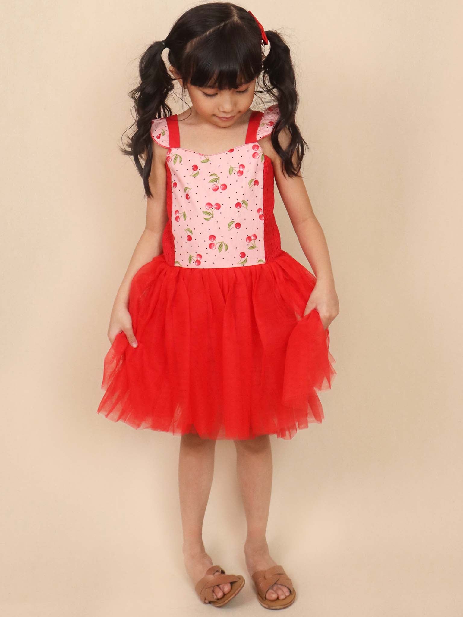 A child in a twirl-approved, red and white floral dress stands against a beige background, showcasing the charm and comfort of the Cherry Ripe Belle Tutu Dress by Retro Cherry.