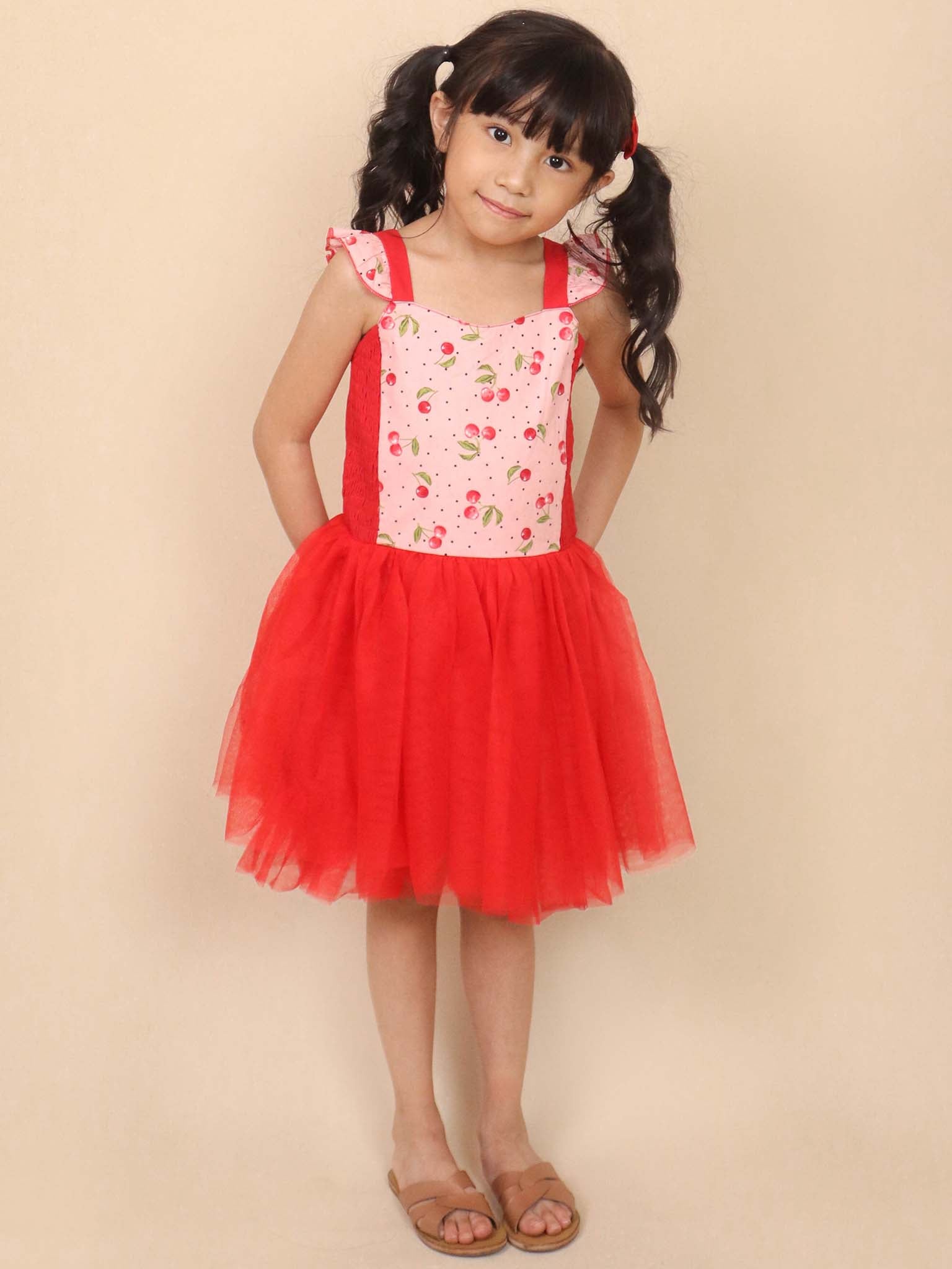 A young girl stands against a plain background, wearing the Cherry Ripe Belle Tutu Dress by Retro Cherry. The red and white dress features a charming cherry pattern with a soft cotton bodice, complemented by her sandals and hair tied with red bows, making it perfectly twirl-approved.