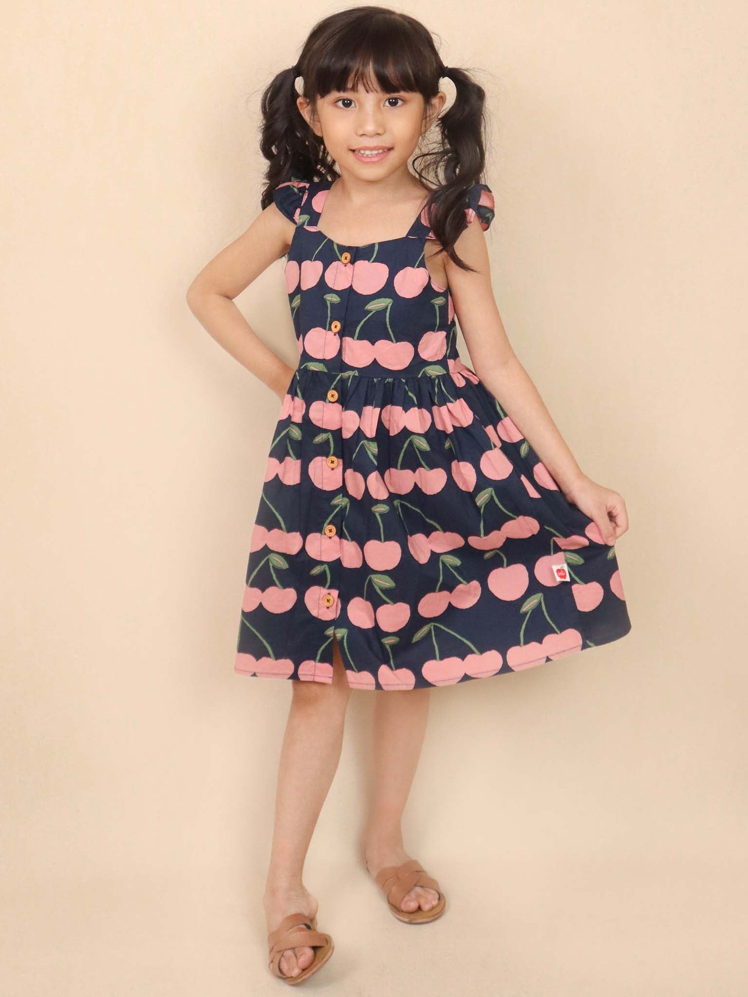 A young girl in a Navy Cherry Calista Dress, featuring flutter ruffles and pink cherry designs, paired with sandals, poses against a beige background.