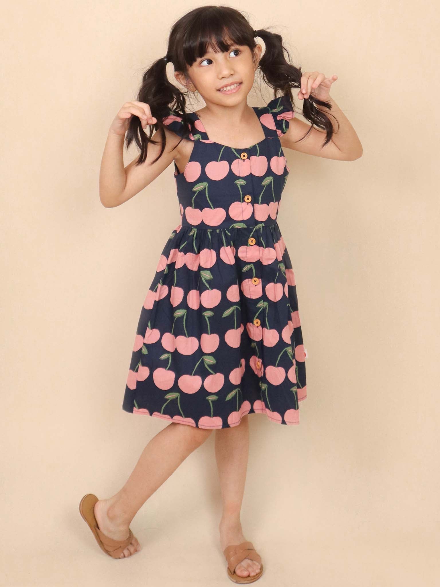 A young girl wearing the Navy Cherry Calista Dress smiles and holds her pigtails against a beige background, showcasing the playful flutter ruffle that adds charm to her outfit.