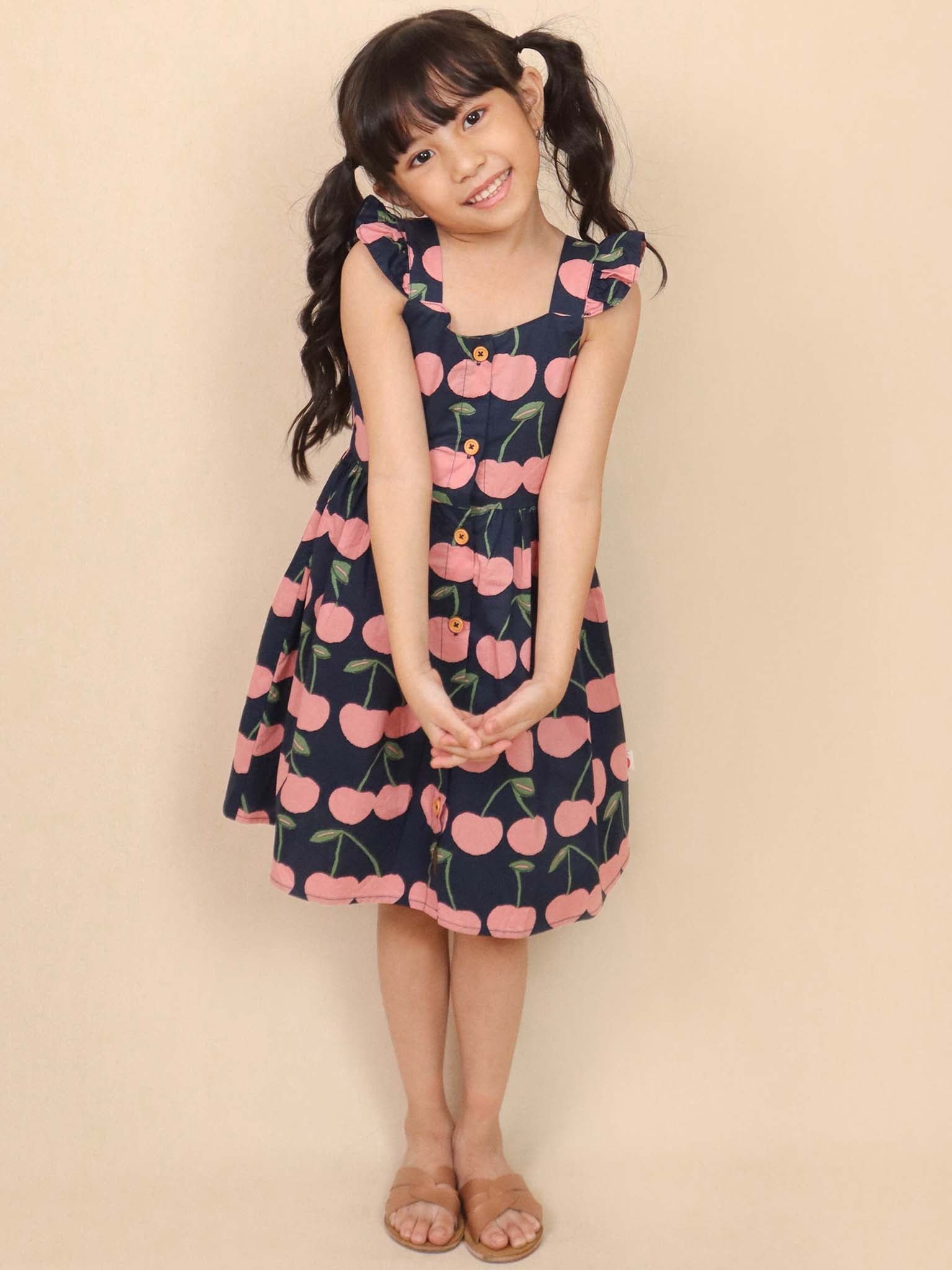 A young girl stands smiling in a Navy Cherry Calista Dress, paired with sandals.