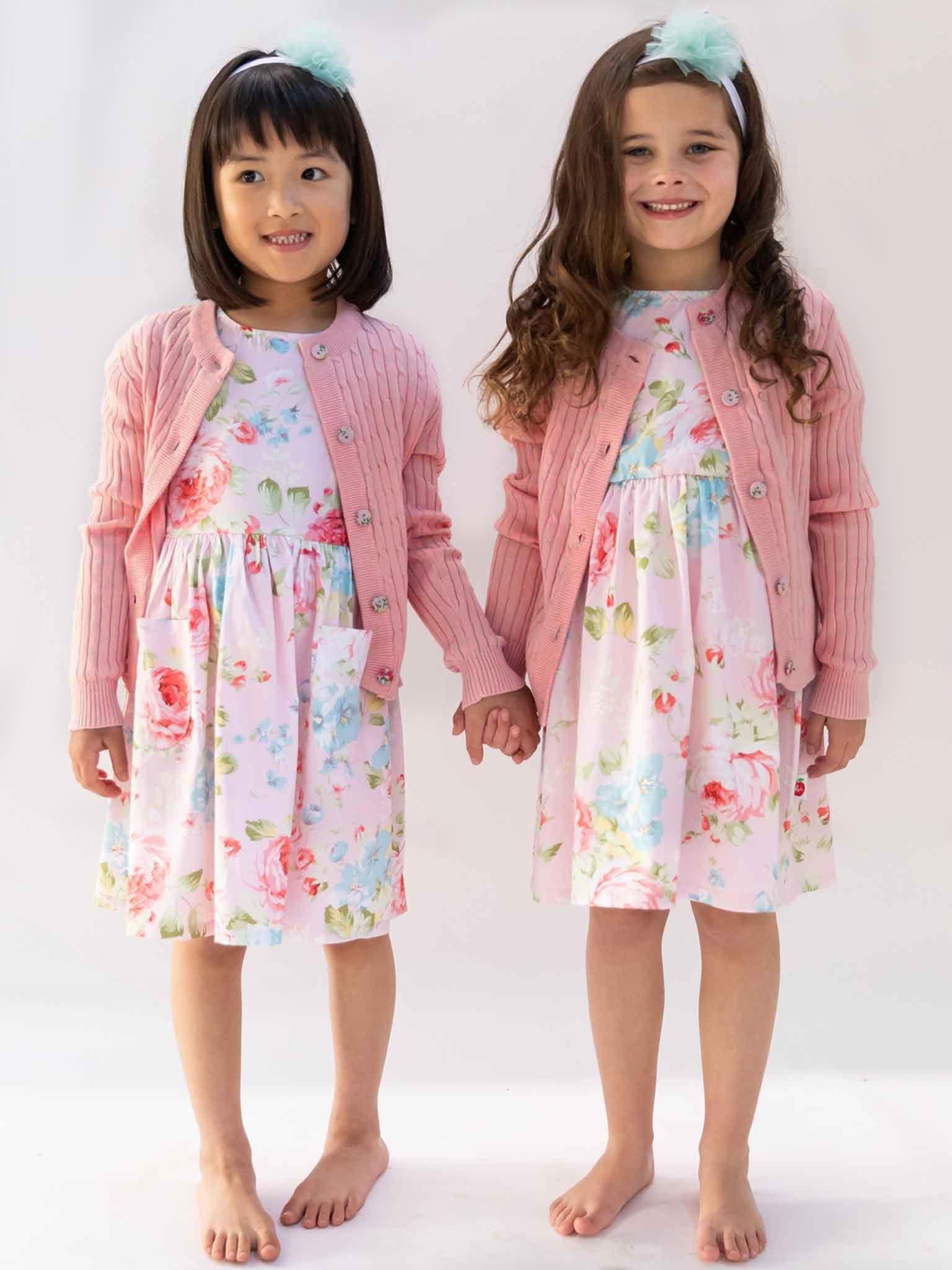 Two girls in matching floral dresses and Knitwear's pink Maxine Icing Cardigans stand barefoot, holding hands and smiling. The 100% cotton cable knit adds cozy charm, while fabric-covered buttons complete their adorable ensemble.