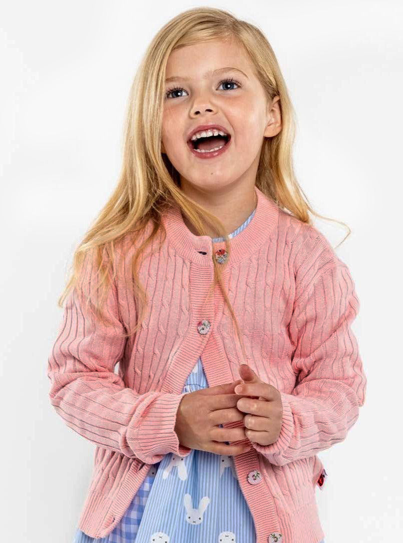 A young girl with long blonde hair smiles wearing a Knitwear Maxine Pink Icing Cardigan made of 100% cotton cable knit. Her blue dress, embellished with white patterns and fabric-covered buttons, enhances her charm as she gazes slightly upward against a plain backdrop.