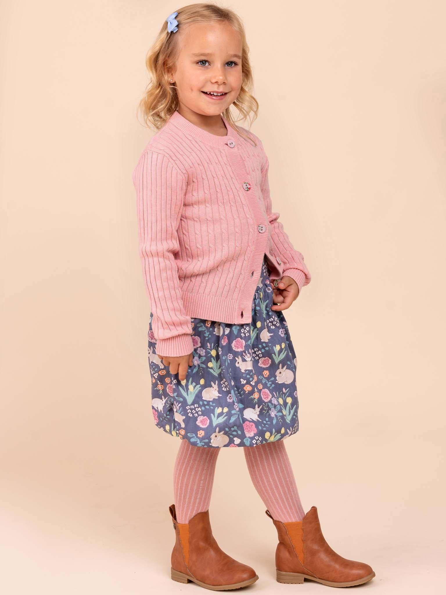 A young girl smiles wearing a Knitwear Maxine Pink Icing Cardigan over a floral dress. She dons pink tights, brown boots, a blue bow in her hair, and has fabric-covered buttons for added charm.