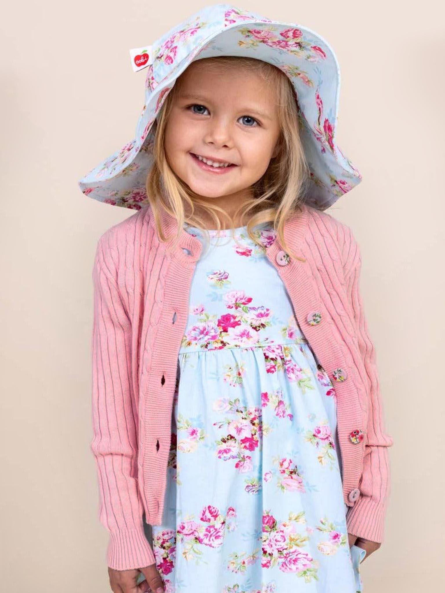 A young girl smiles against a plain background, wearing a Knitwear Maxine Pink Icing Cardigan with fabric-covered buttons, paired with a floral dress and matching sun hat.