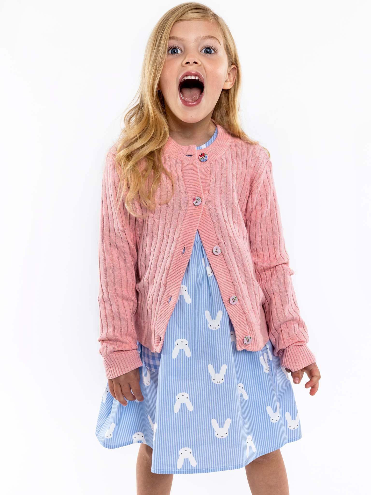 A young girl with long blonde hair wears a Knitwear Maxine Pink Icing Cardigan in 100% cotton cable knit and a blue dress with rabbit prints, her mouth open in surprise; each detail is highlighted by fabric-covered buttons.