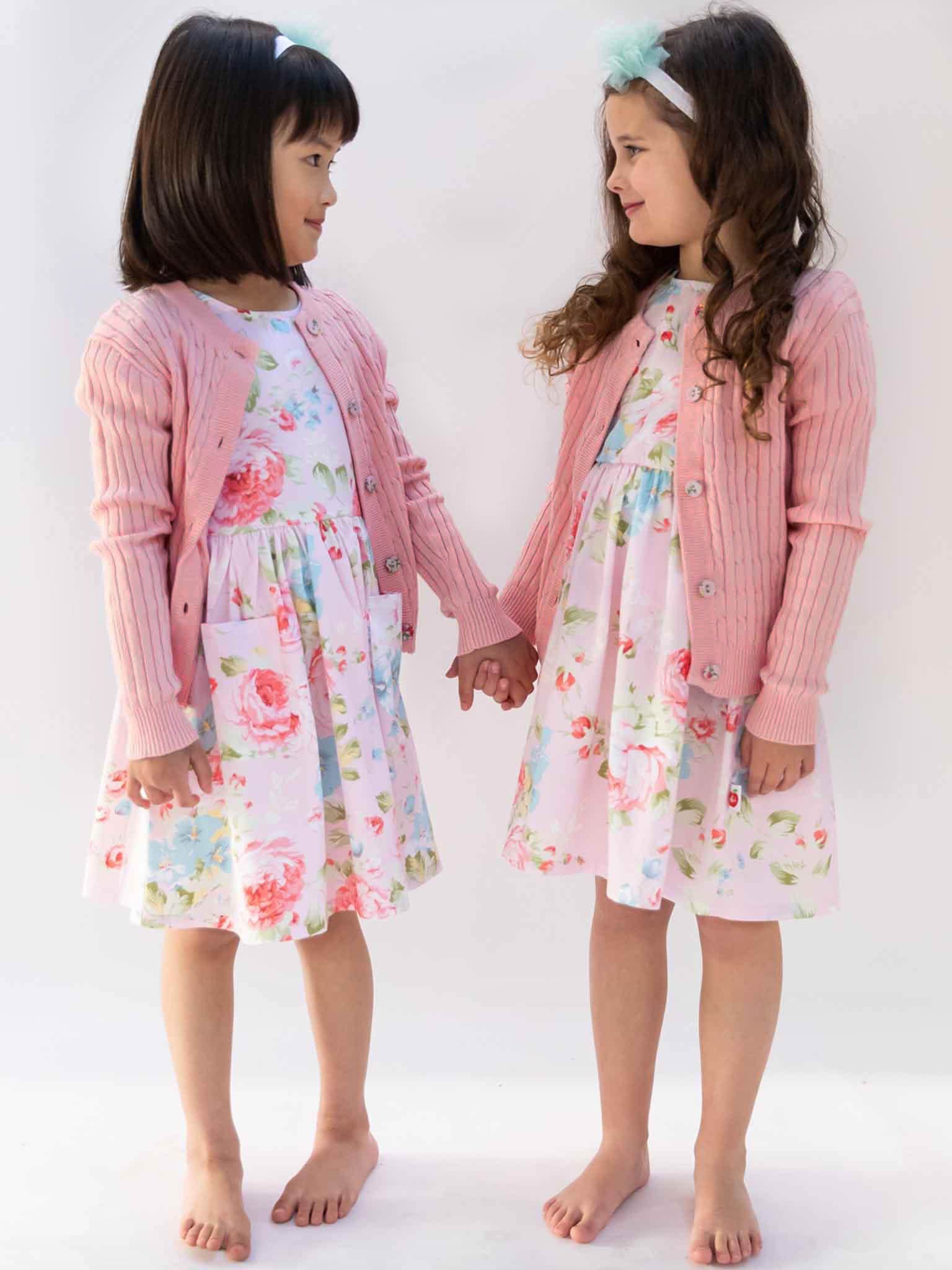 Two young girls stand barefoot, facing each other and holding hands, wearing matching floral dresses with Knitwear's Maxine Pink Icing Cardigans featuring fabric-covered buttons, accented by headbands in their hair.