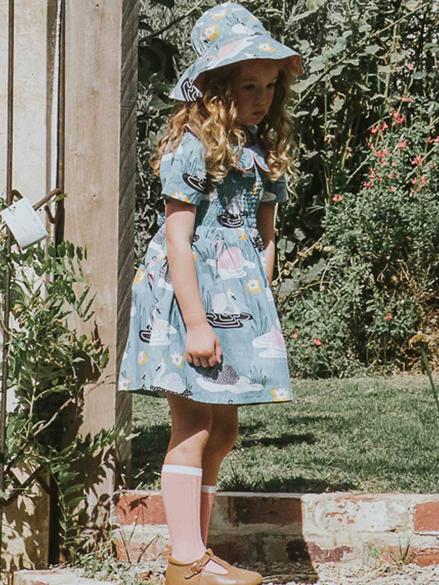 A young girl with wavy hair stands outside wearing the Swan Song Bucket Hat by Swan Song, along with a matching blue dress adorned with floral patterns, pink socks, and brown shoes. Her 100% cotton outfit complements the wooden post and garden greenery nearby.