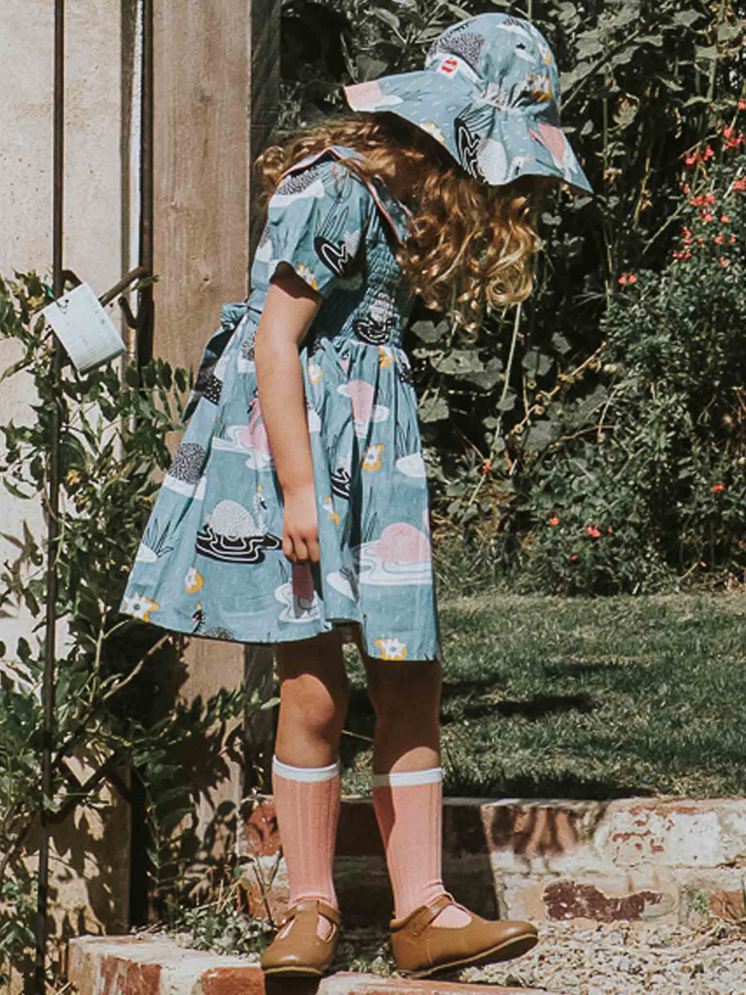 A young girl in a fitted blue printed dress and the Swan Song Bucket Hat from Swan Song stands outdoors, looking down. She is wearing pink knee-high socks and brown shoes against a backdrop of greenery and a brick pathway.