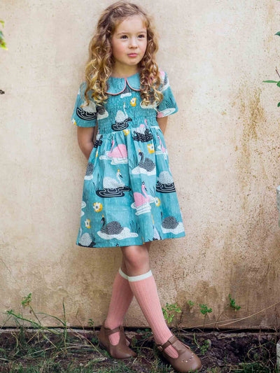 A young girl with curly hair standing outdoors against a textured wall, dressed in the Swan Song Rebecca Dress featuring the vintage Swan Song print. She is paired with pink knee-high socks and brown shoes.