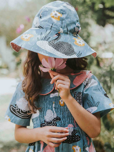 A young girl wearing a Swan Song 100% cotton floral-patterned hat and matching dress holds a pink flower to her nose while standing outdoors.