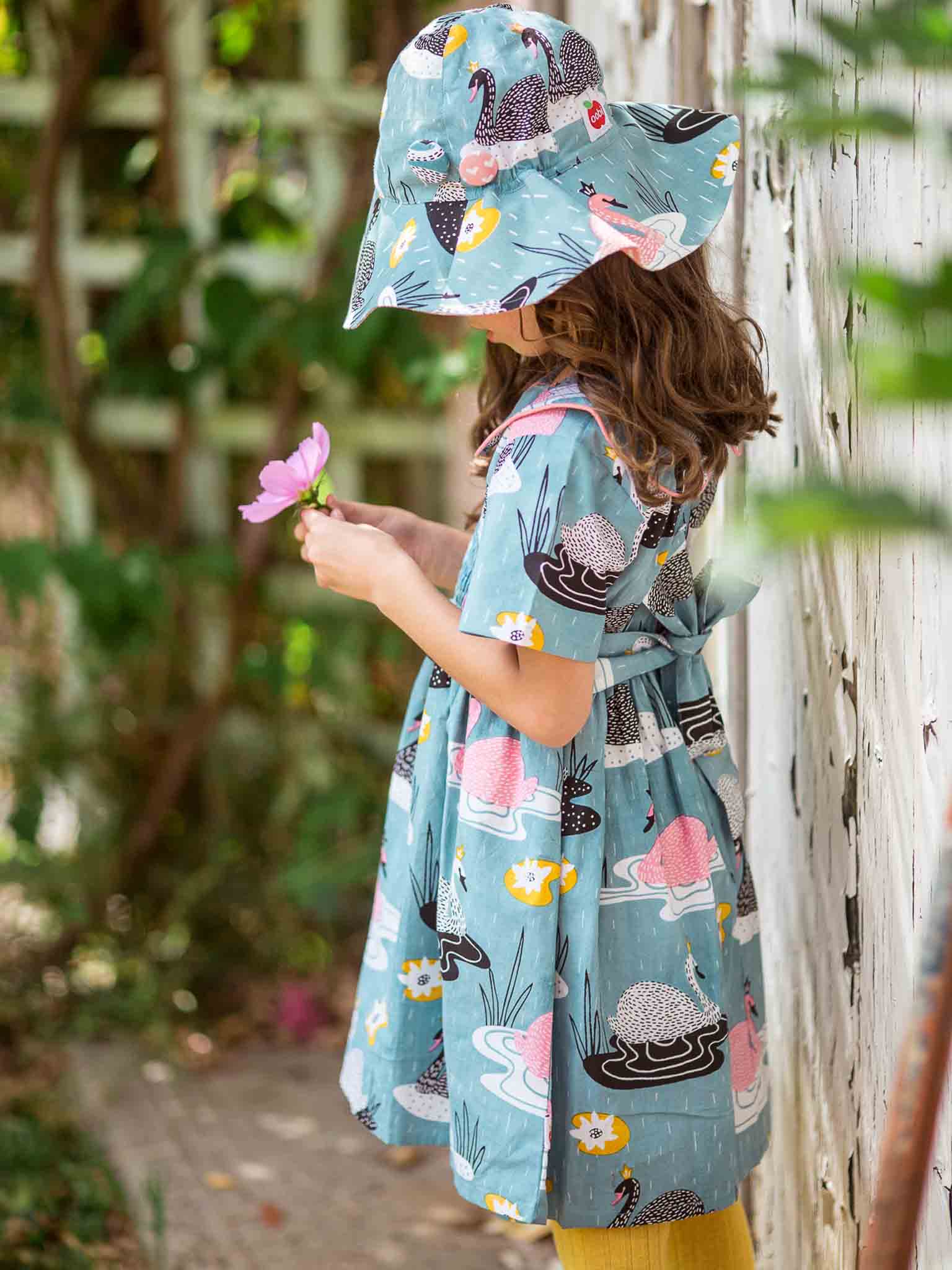 A young girl in a blue, 100% cotton dress and a matching Swan Song Bucket Hat with animal prints stands outdoors, holding a pink flower. She is facing a white fence with green foliage in the background.