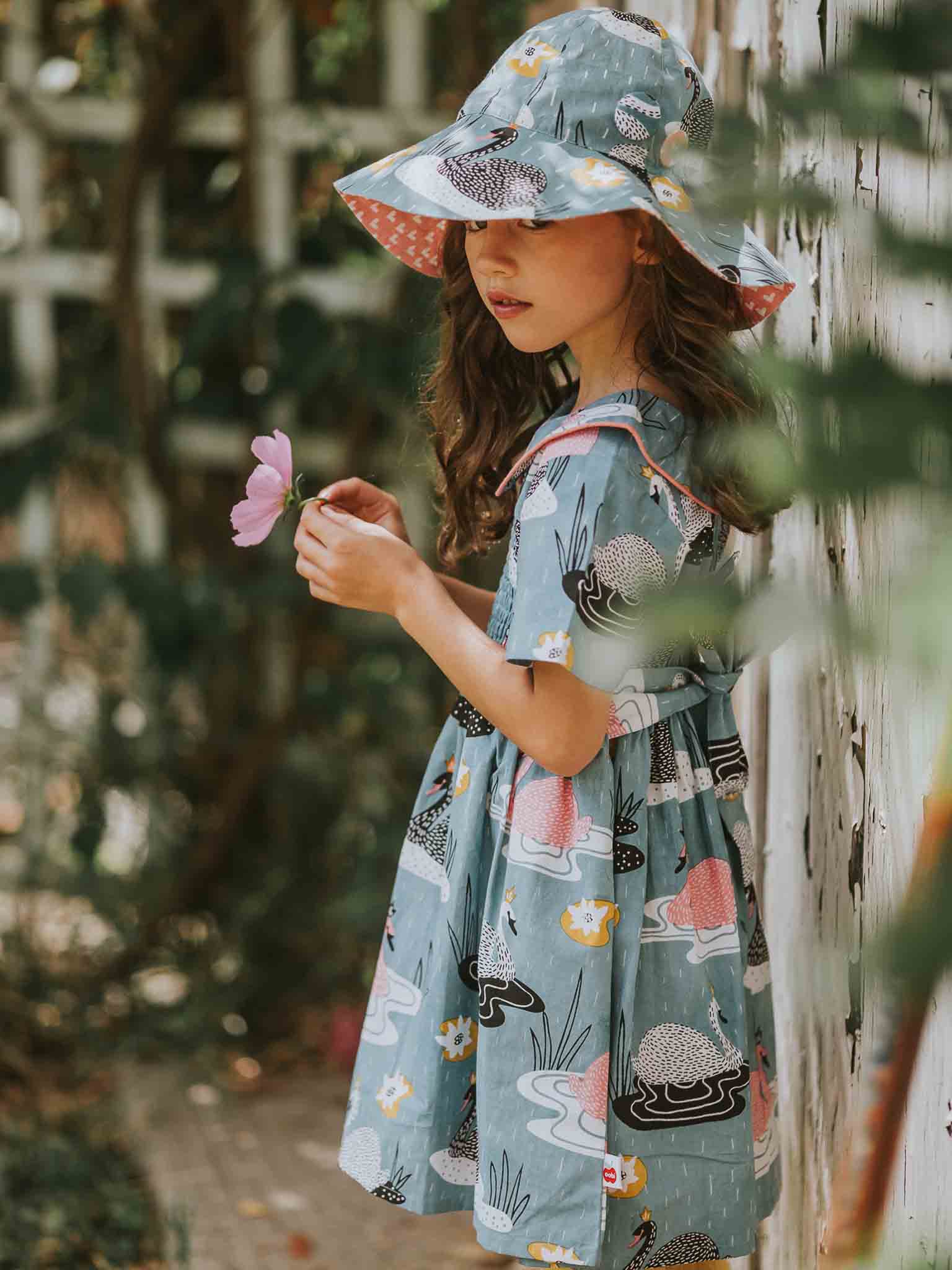 A young girl stands outdoors in a blue patterned dress with a fitted design, accessorized by the Swan Song Bucket Hat—a matching 100% cotton hat from the Swan Song brand. She holds a pink flower while looking away.