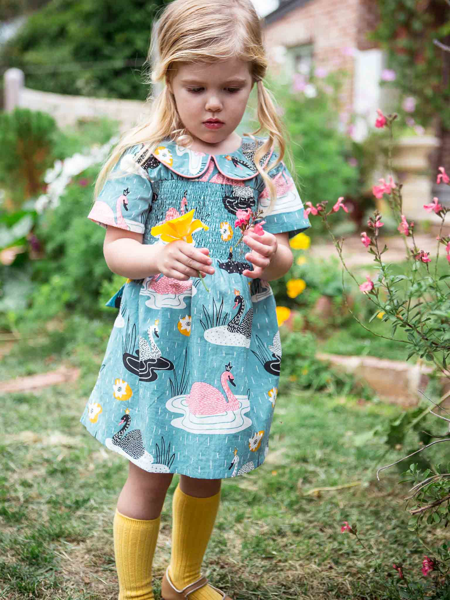 A young girl in the Swan Song Rebecca Dress, featuring a Peter Pan collar and vintage Swan Song print, stands in a garden with yellow socks peeking out as she holds a yellow flower and looks down.