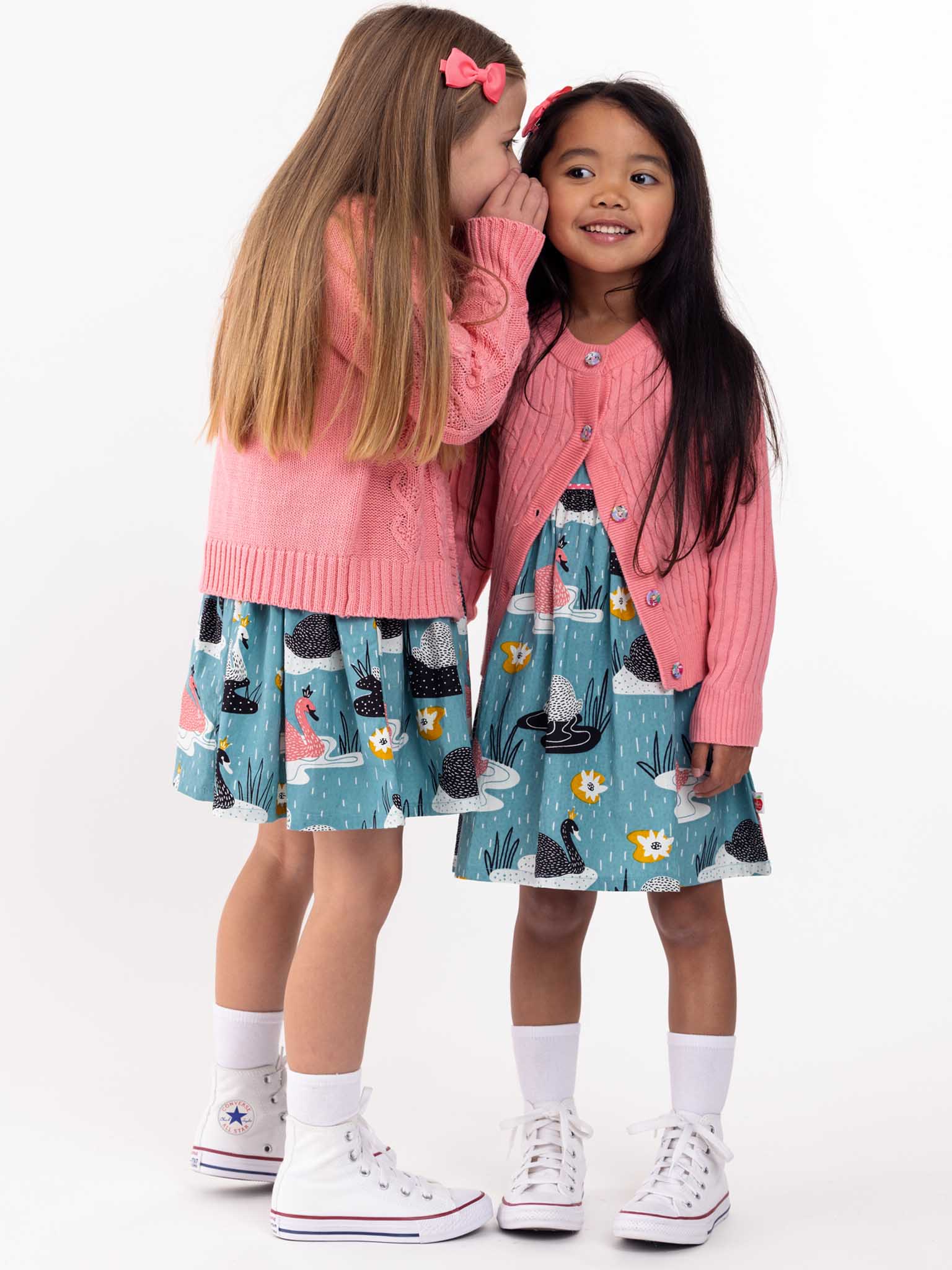 Two young girls in matching Knitwear Willow Geranium Pink Cotton Cardigans whispering secrets, standing against a white background.