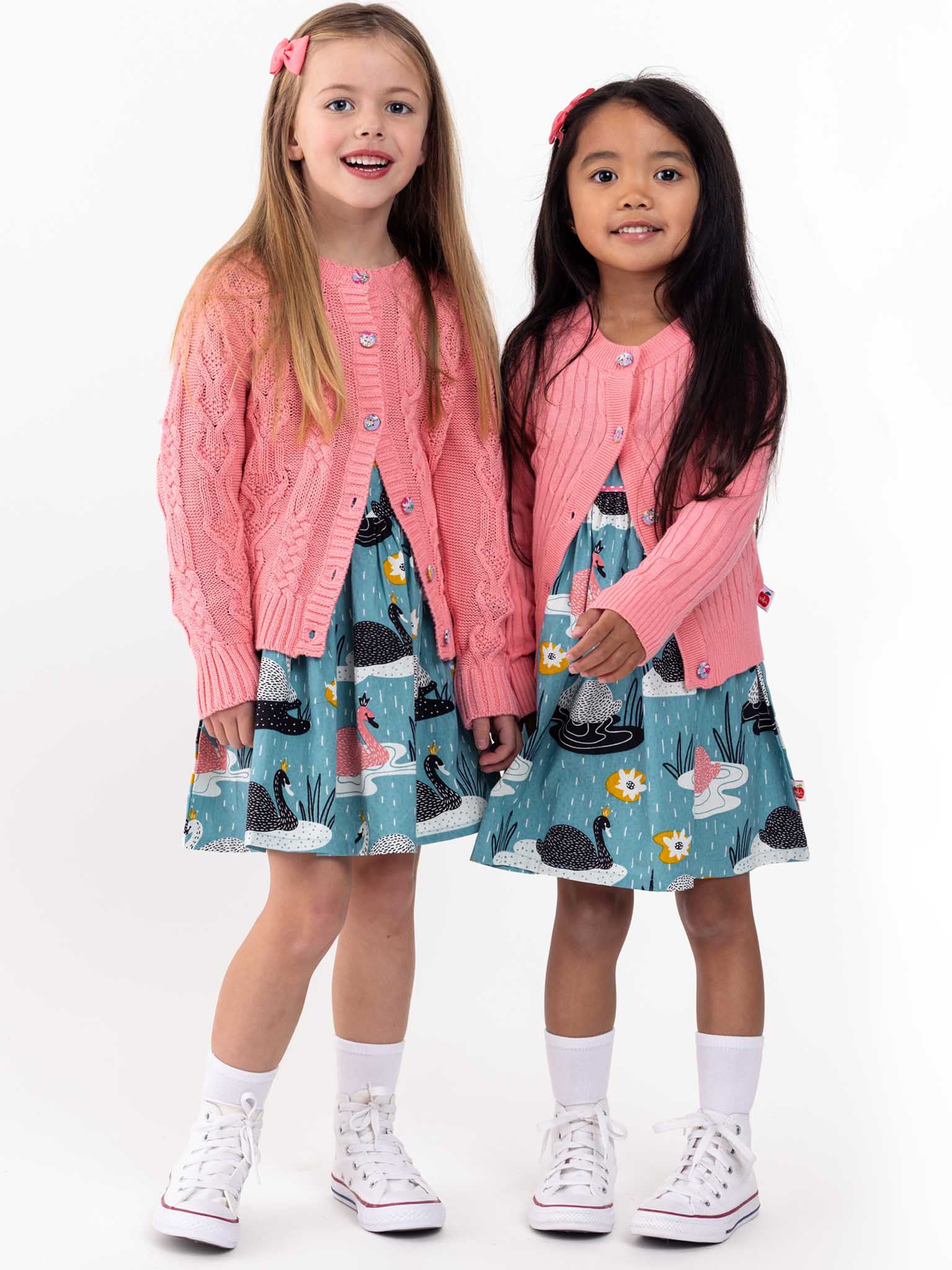 Two young girls in matching pink Knitwear Willow Geranium Pink Cotton Cardigans and floral dresses with sneakers, standing and smiling against a white background.