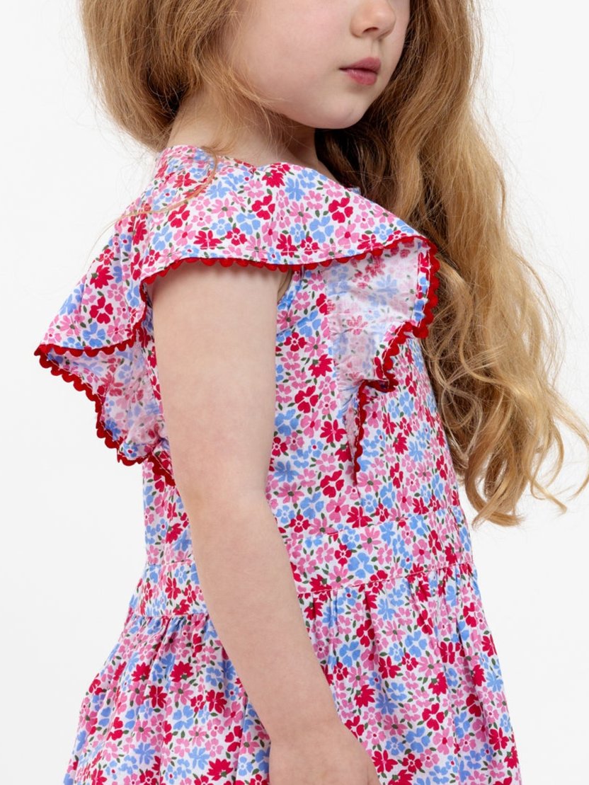 A young girl with long hair is wearing the Cottage Flowers Red Cottage Flowers Lace Edge Dress, which features colorful, floral patterns and lace-trimmed, ruffled sleeves, as she stands facing sideways.