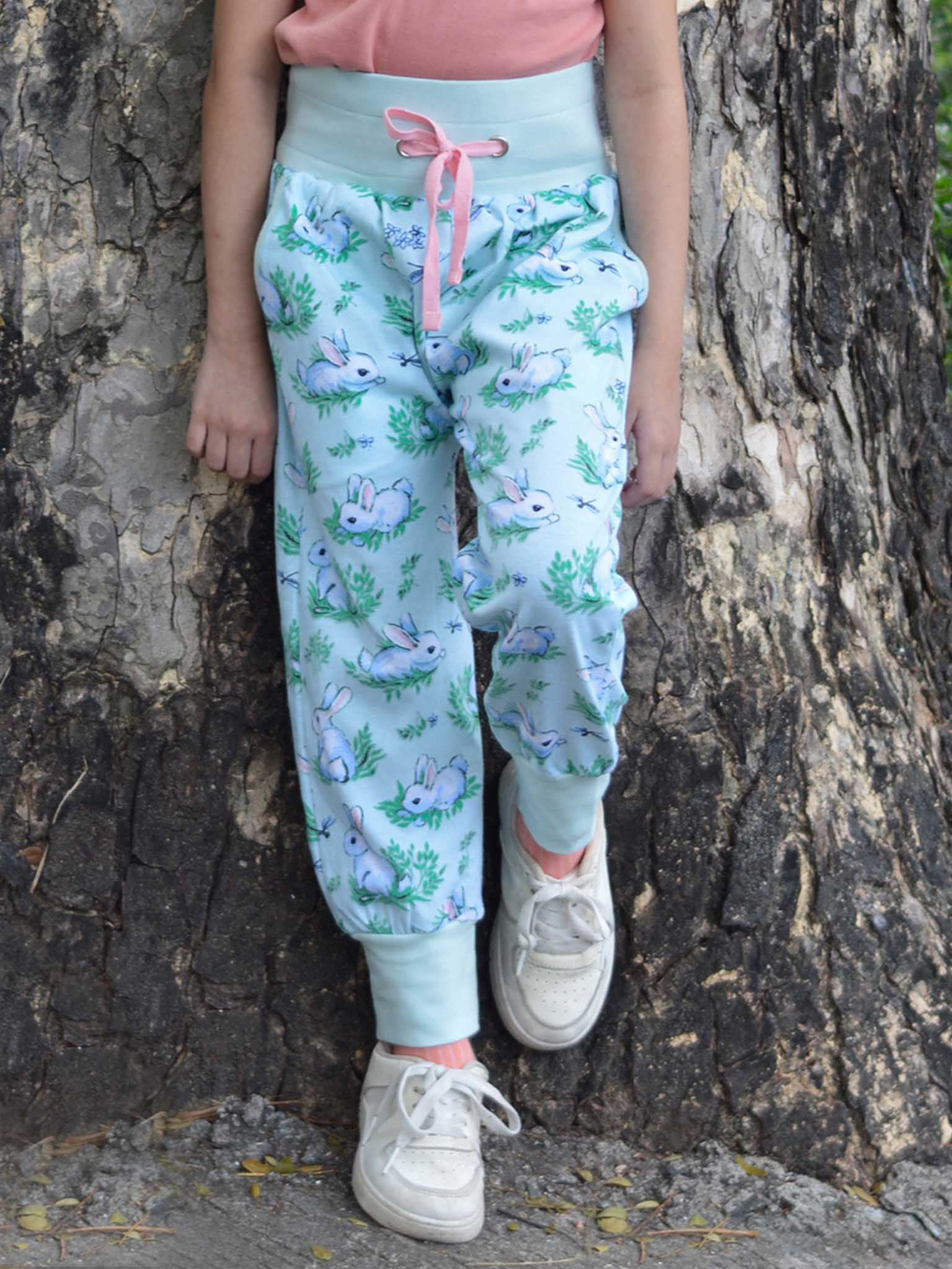 A child in Blue Bun-Buns Soft Jersey Pants with an elastic waist and bunny print, a pink top, and white sneakers stands by a tree. These comfy pants from Bun-Buns are stylish and popular for playful days.