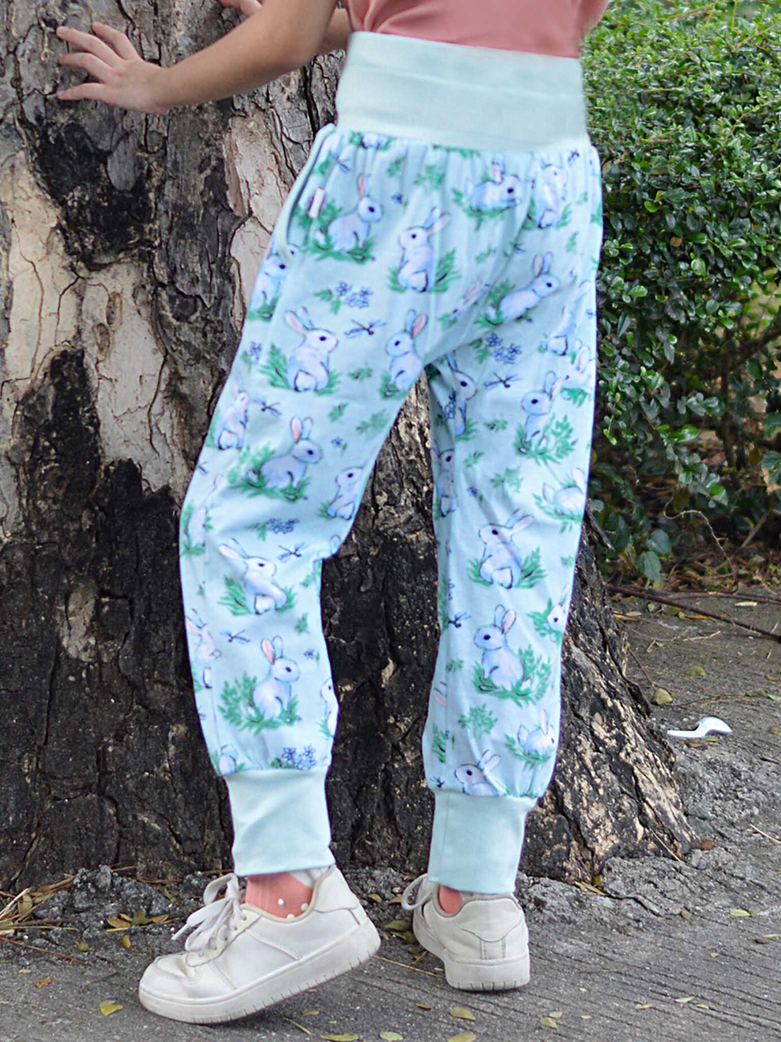 A child stands outdoors near a tree in Bun-Buns' Blue Bun-Buns Soft Jersey Pants, featuring an elastic waist for added comfort and decorated with bunny prints, making these pants a best seller.