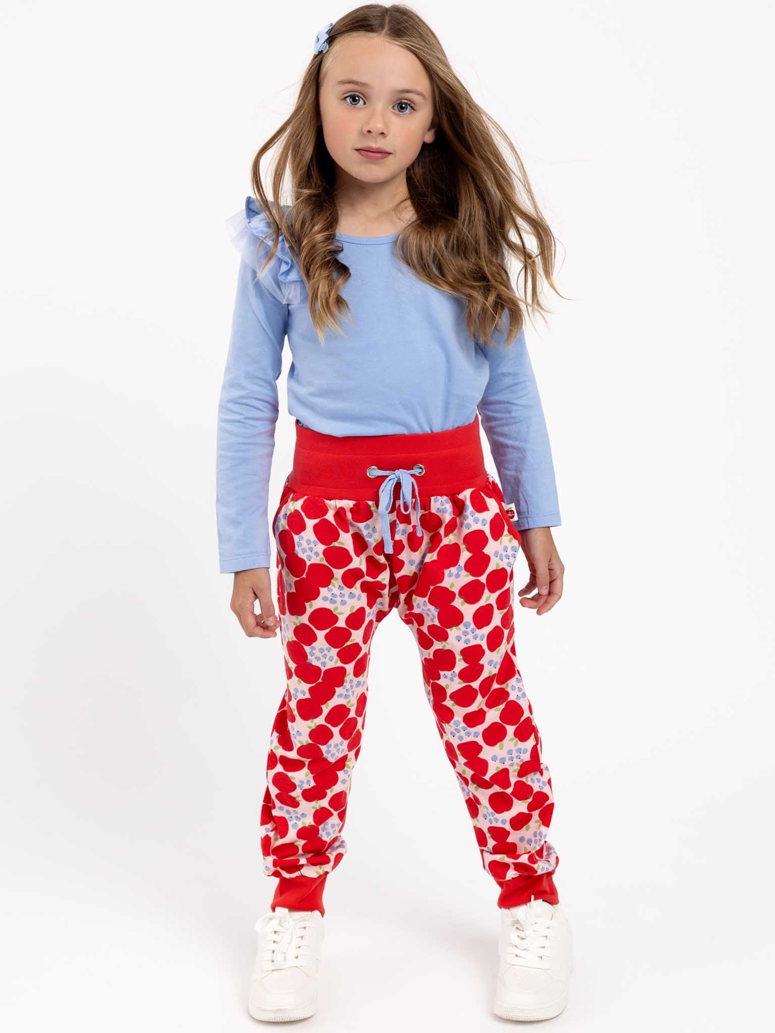 A child stands against a white background wearing the Essentials Sweetheart Blue Tee - L/S, red and white heart-patterned pants, and white sneakers.