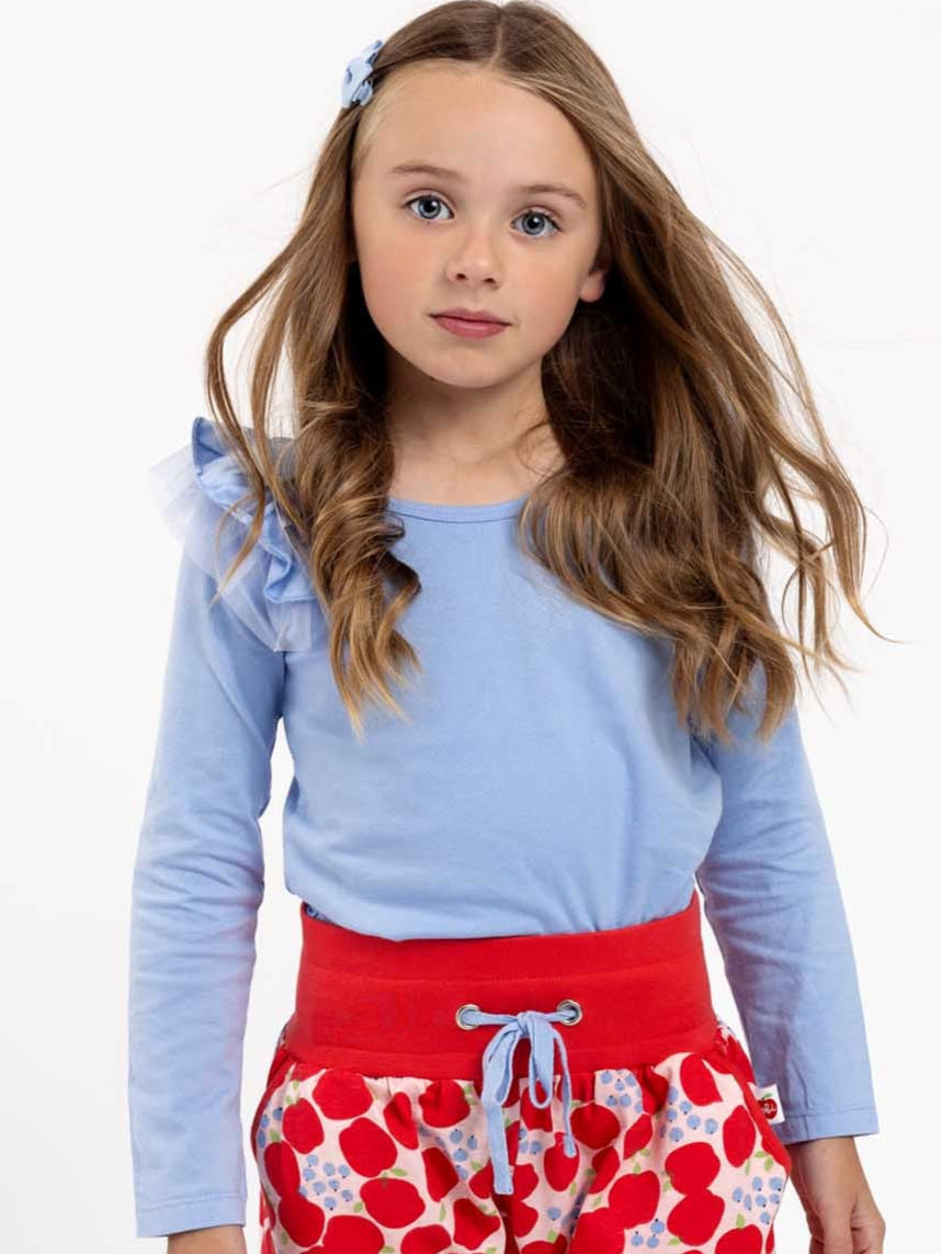 A young girl with long hair wears the Essentials Sweetheart Blue Tee - L/S and a red skirt with heart patterns, standing against a plain white background.