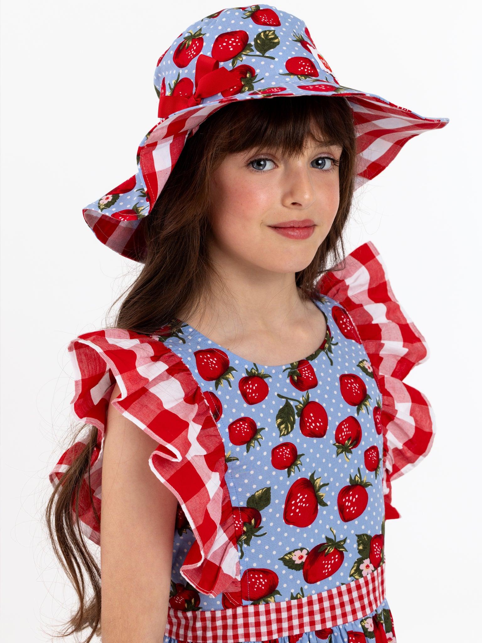 A little girl wearing a Strawberry Fields Jayne dress with a festive strawberry print hat.