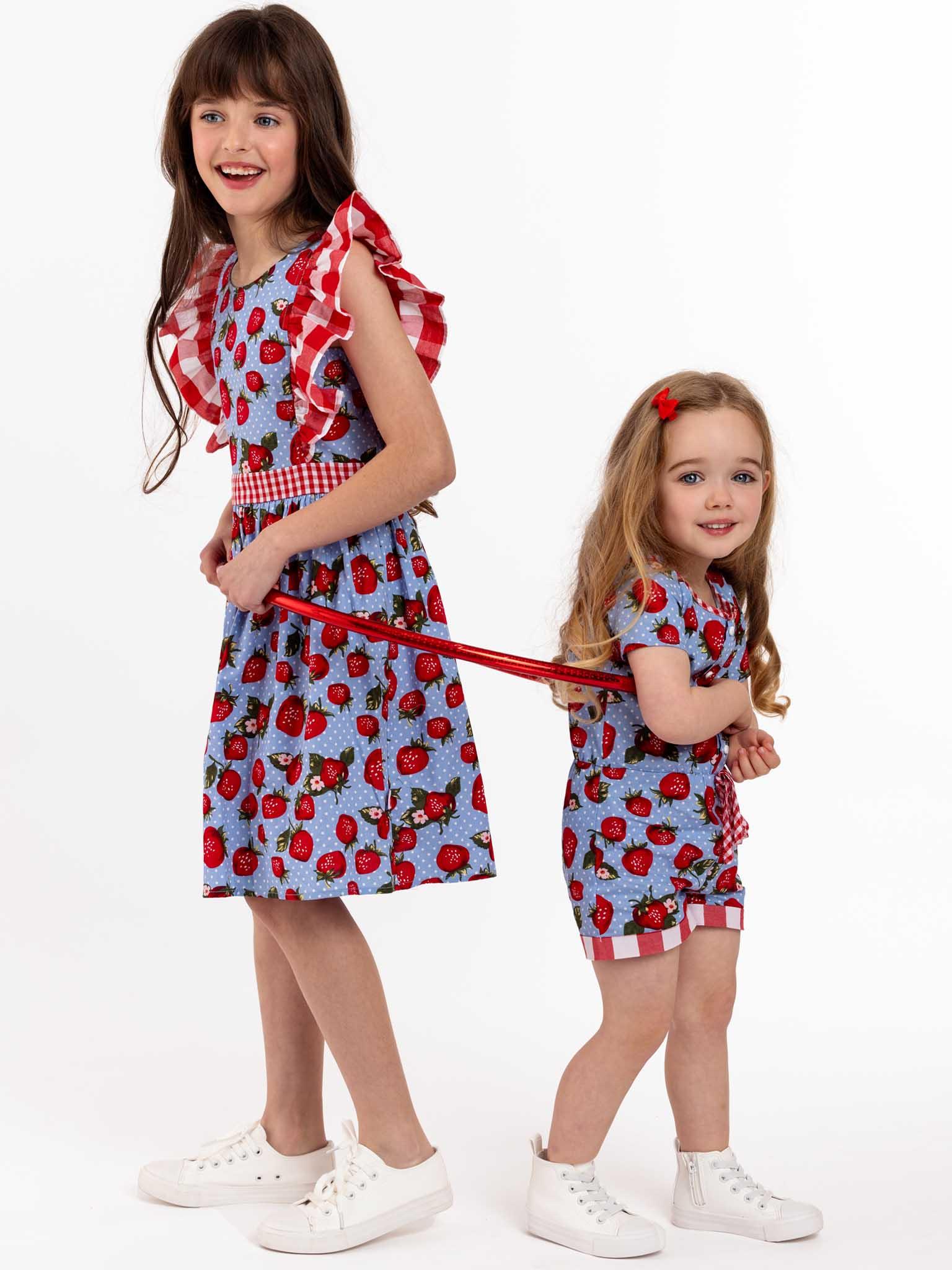 Two little girls in festive dresses are standing next to each other holding a leash, wearing Strawberry Fields Jayne Dresses from the brand Strawberry Fields.