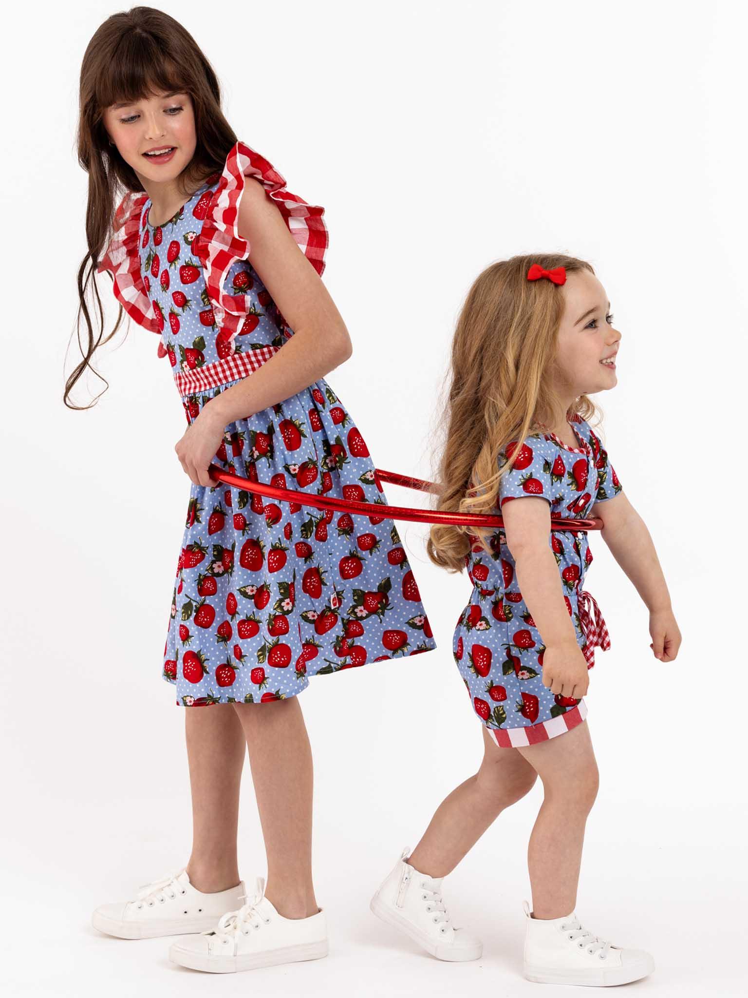 Two little girls in Strawberry Fields Jayne Dresses are walking with a leash in front of a white background.