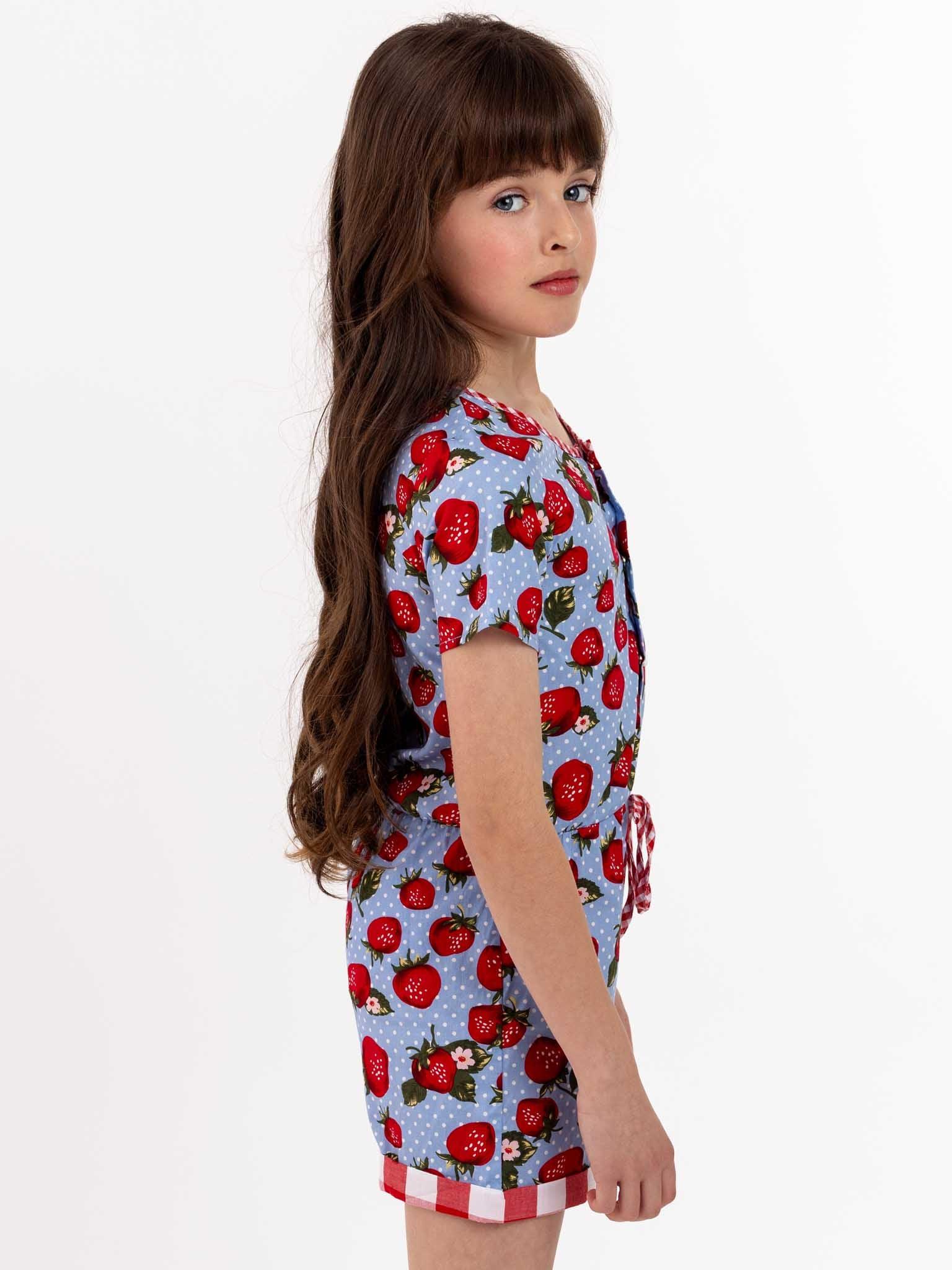 A little girl wearing a Strawberry Fields Maisy Playsuit, an easy-to-wear blue and red strawberry print romper with press studs.