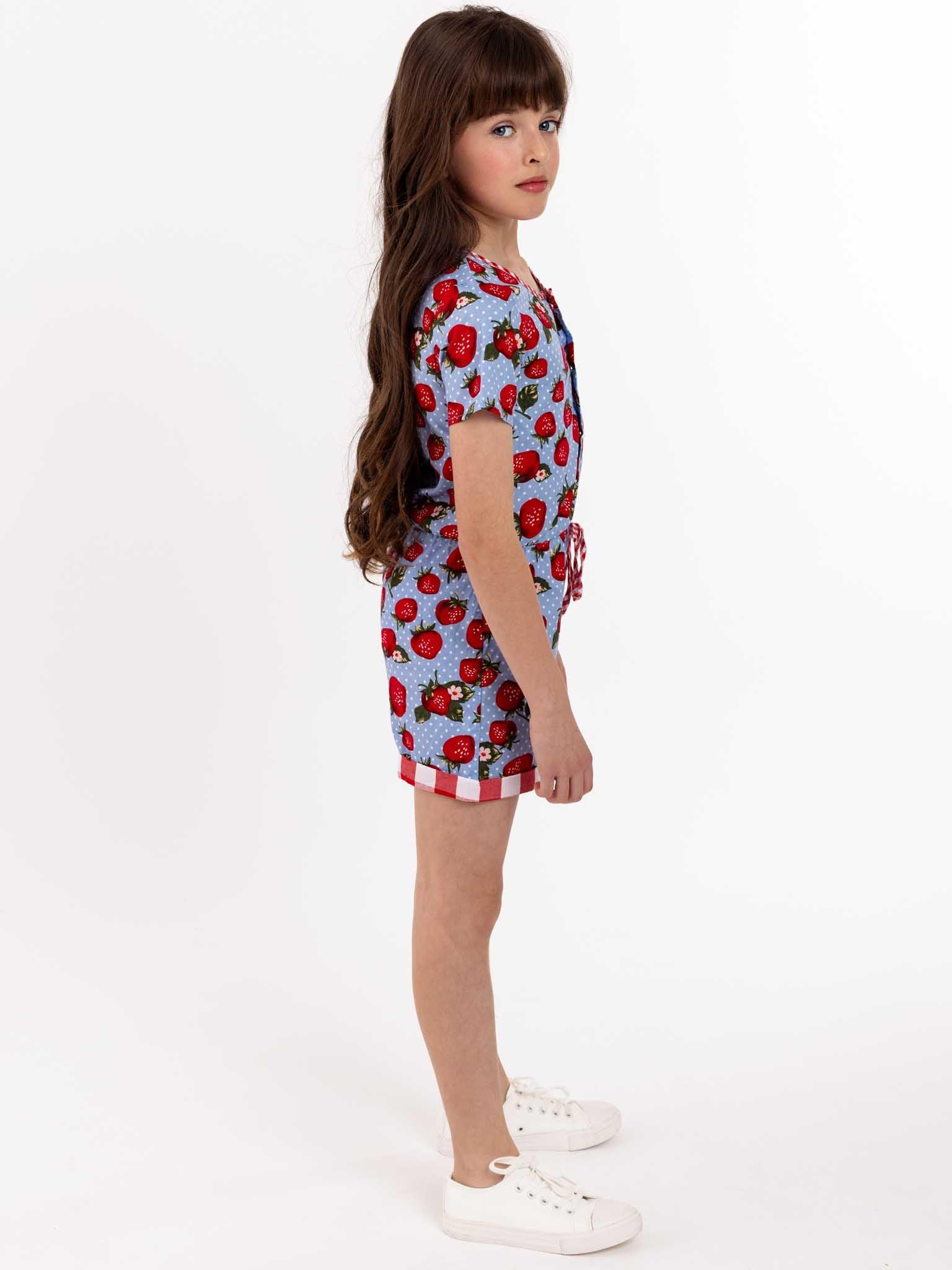 A little girl in a Strawberry Fields Maisy Playsuit, an easy-to-wear blue and red strawberry print romper with press studs.