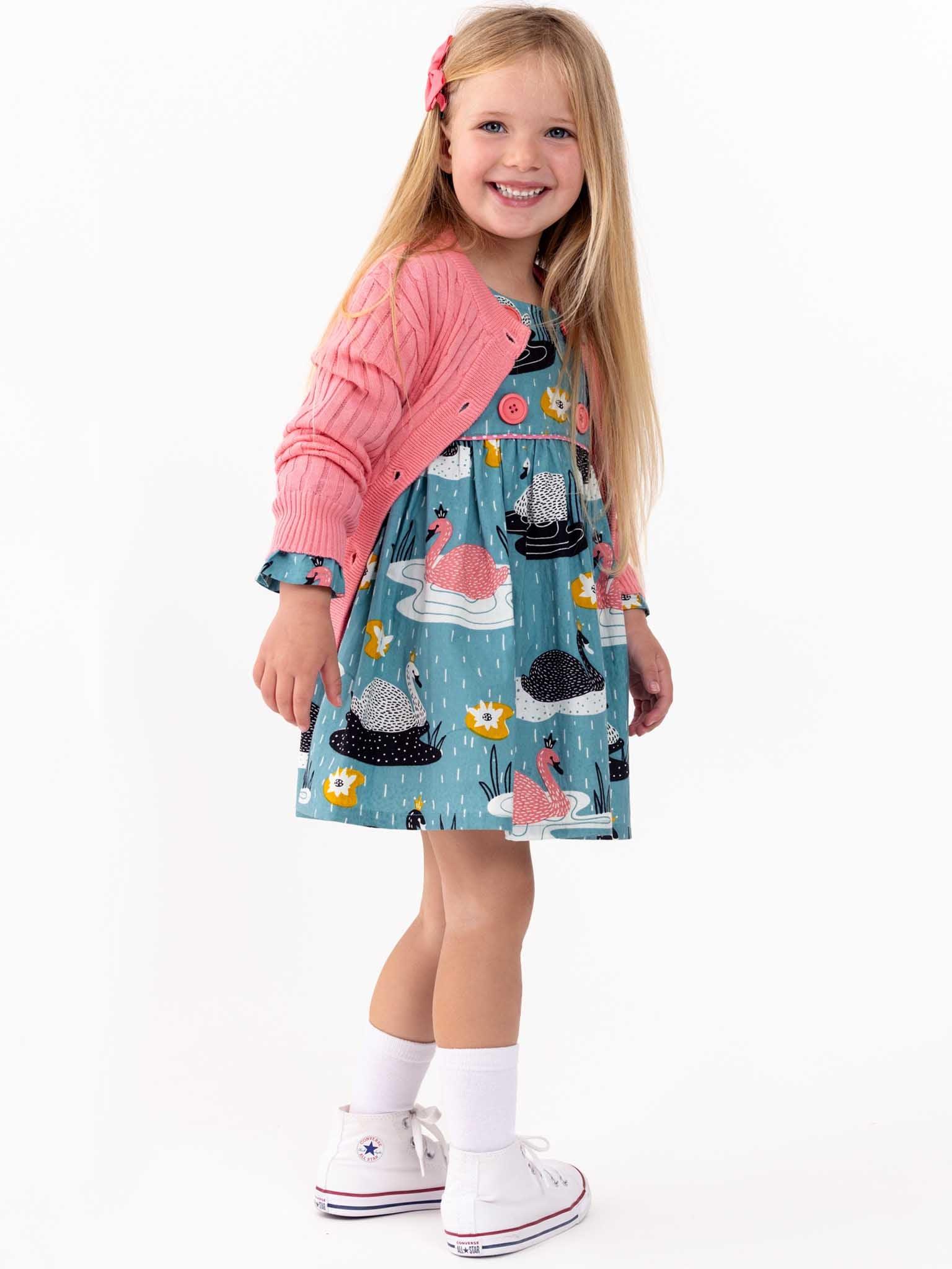 A young girl smiling at the camera, dressed in a colorful Swan Song Sage Dress with a pink cardigan and white sneakers.