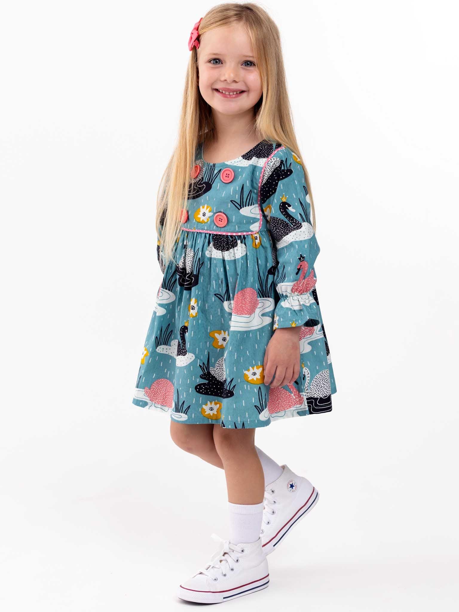 Young girl with long blonde hair smiling, wearing a Swan Song blue dress and white sneakers, standing against a white background.