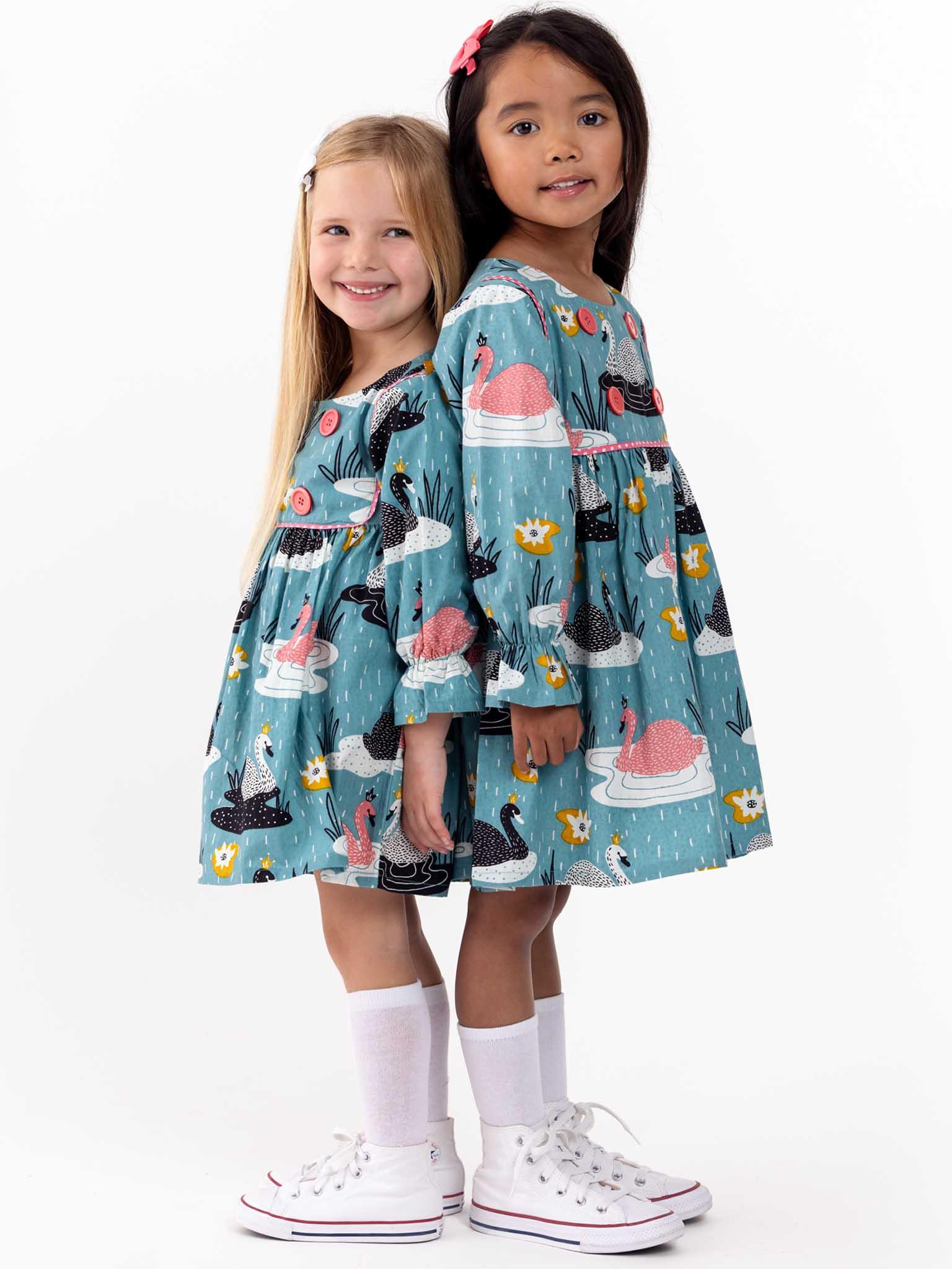 Two young girls wearing matching teal Swan Song Sage Dresses with swan prints, standing side by side, smiling against a white background.