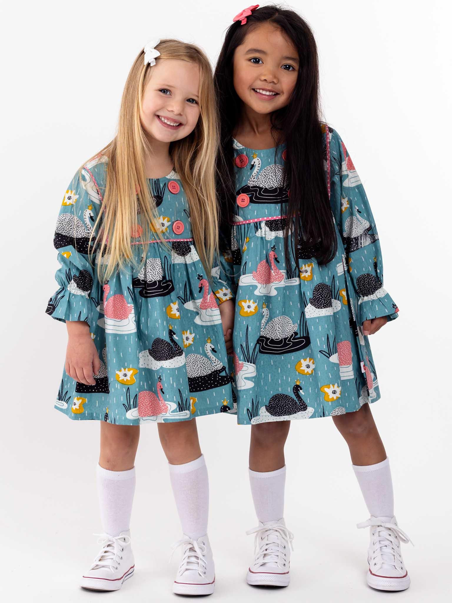 Two young girls wearing matching Swan Song Sage Dresses and white sneakers pose together with a smile, standing against a white background.