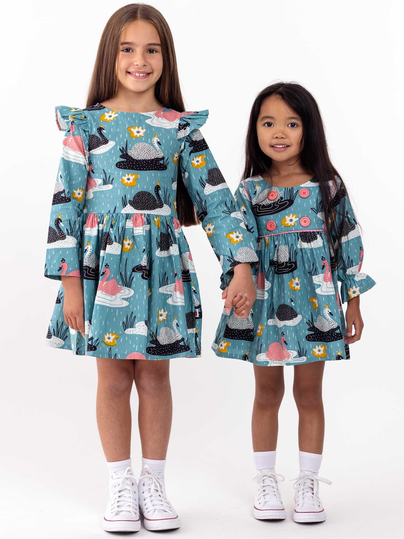 Two young girls smiling and standing side by side, wearing matching Swan Song Sage Dresses with swan prints and white sneakers.